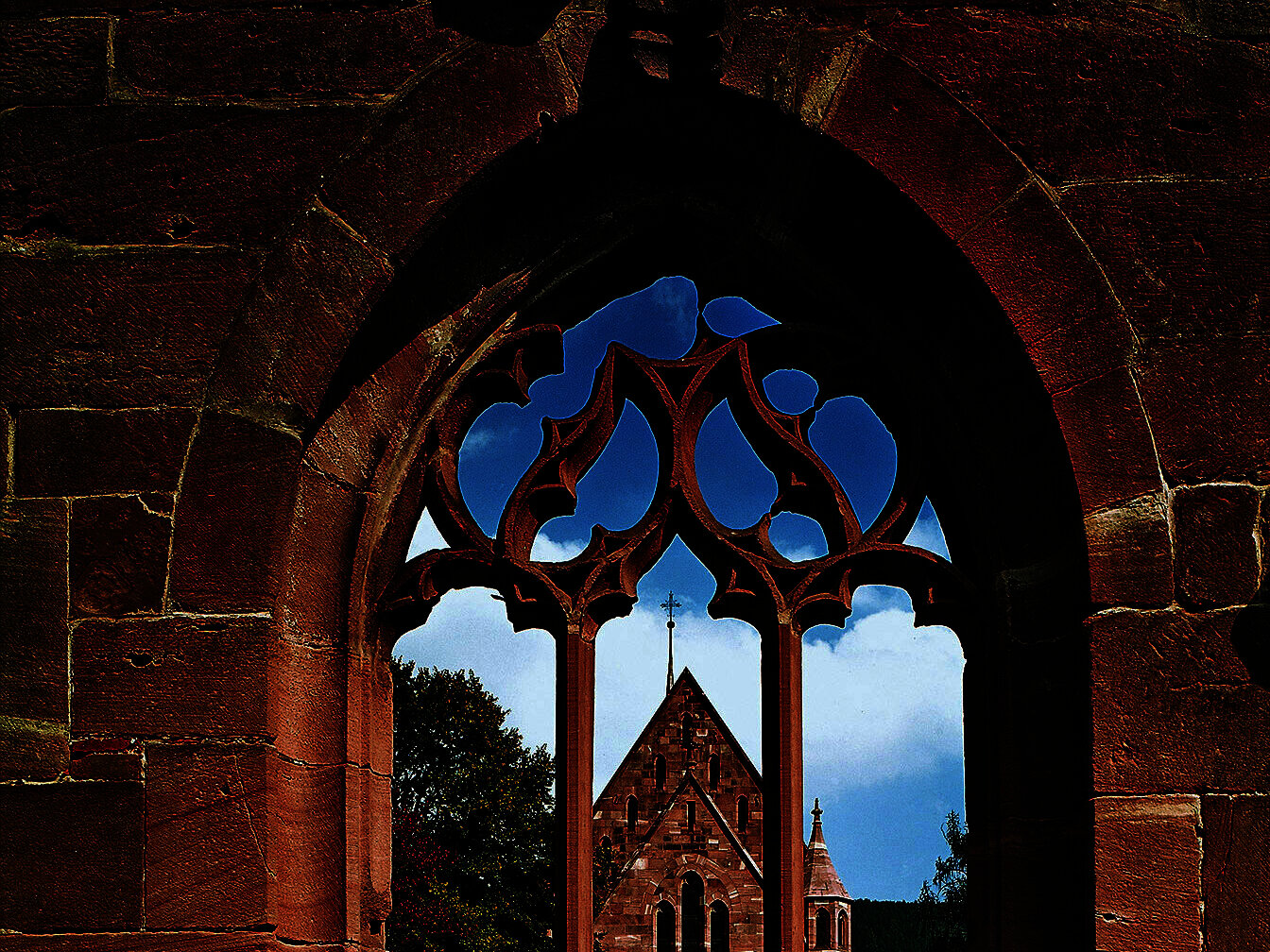 Blick durch einen sandsteinernen gotischen Fensterbogen auf eine ebenfalls sandsteinerne Kapelle.