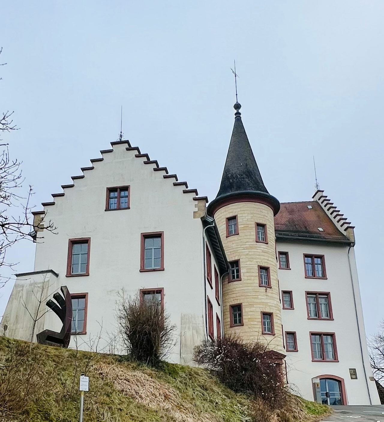 Bergauf fotografiert das Schloss mit Treppengiebel und Rundturm, helle Fassade, rote Fensterrahmen, bewölkter Himmel.