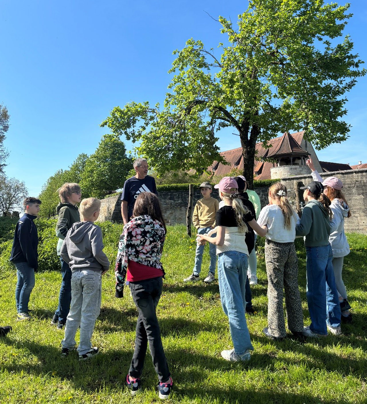 Eine Gruppe von Jugendlichen steht auf einer Wiese neben einem Baum im Kreis und hört einem Vortragenden zu.