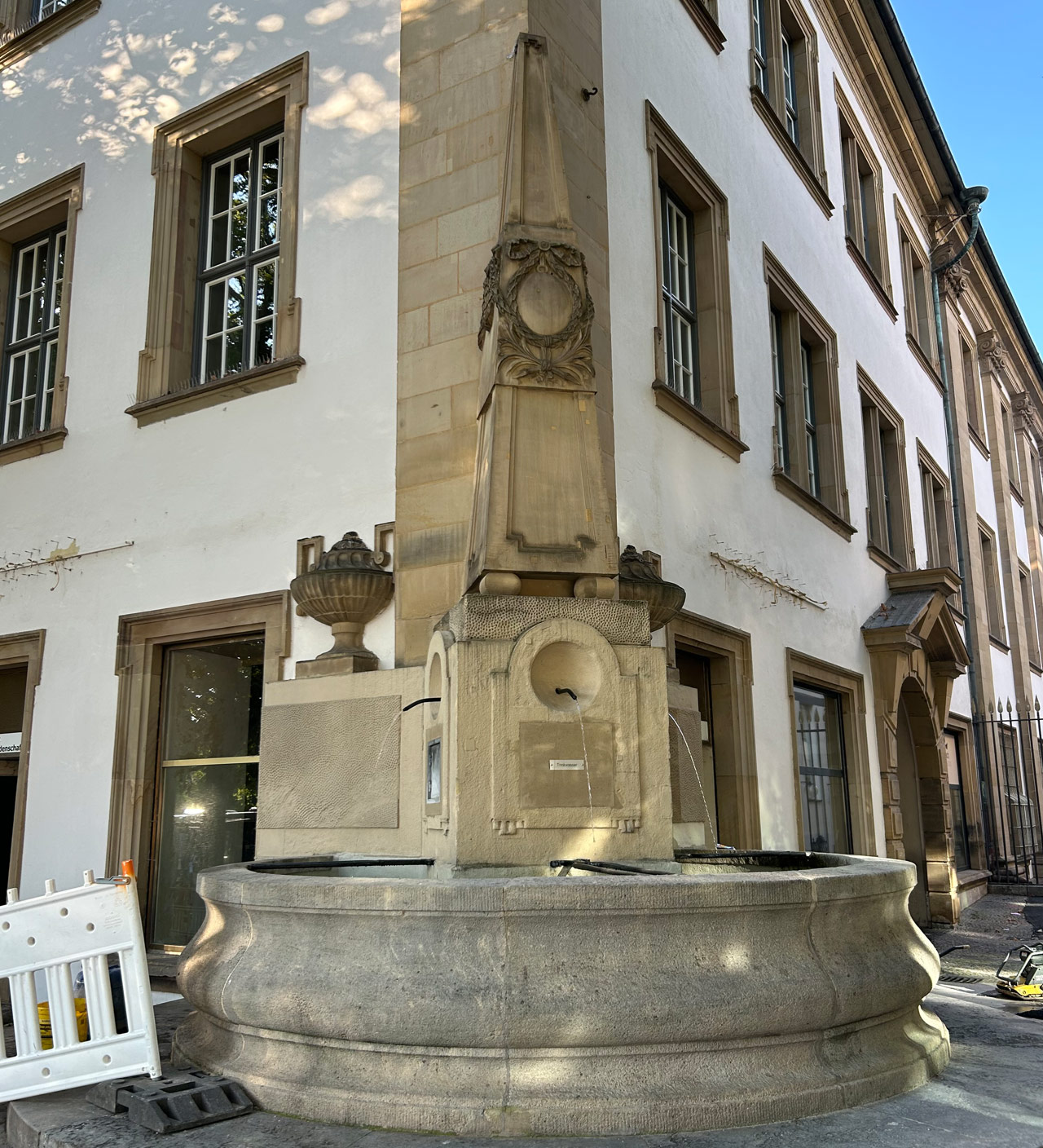 Ein historischer Brunnen, mit steinerner, verzierter Stehle in der Mitte mit drei laufenden Wasserhähnen.