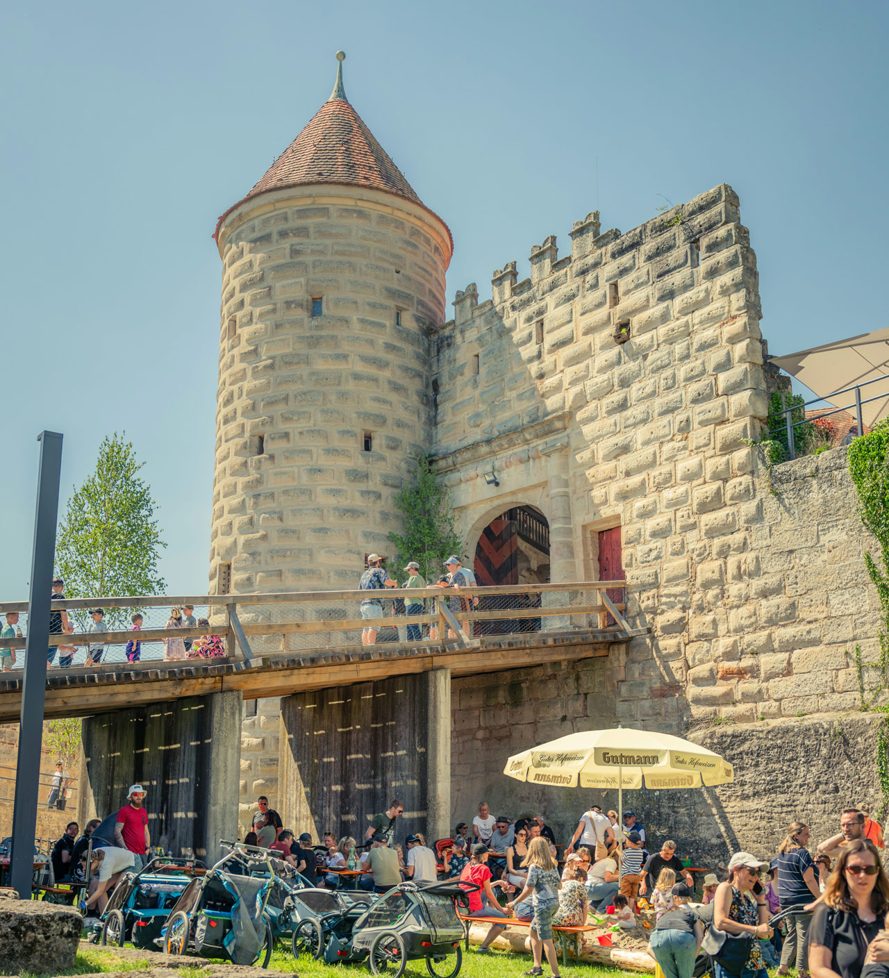 Eine Burg mit Turm und Gemäuer. Sommerliche Stimmung, es halten sich viele Leute auf, vor allem jüngere Menschen mit Kindern. 