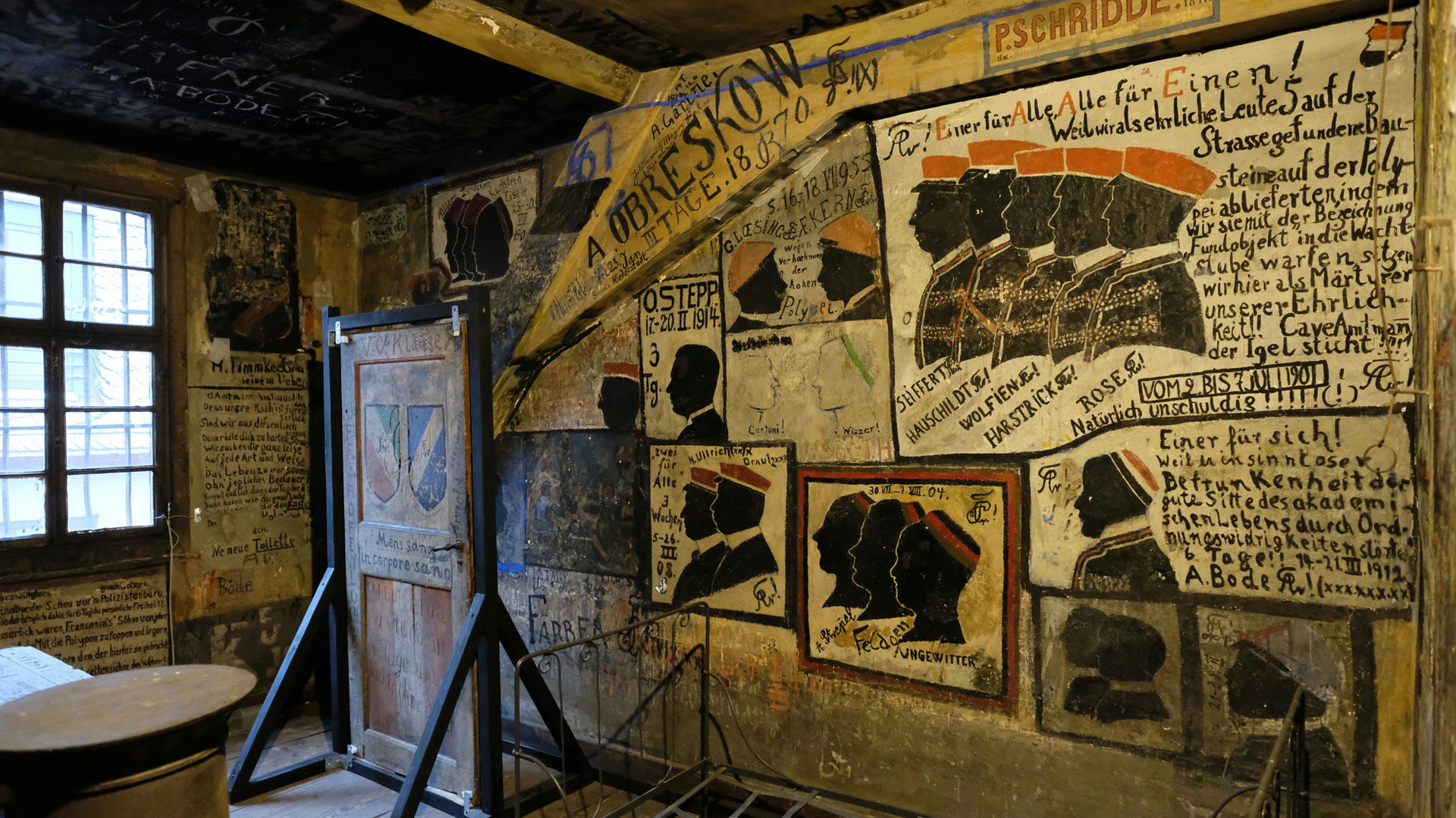 Ein historischer Raum mit Zeichnungen und Malereien an den Wänden. Mehrere Ausstellungsstücke, u. an. eine Tür in einem Gestell.