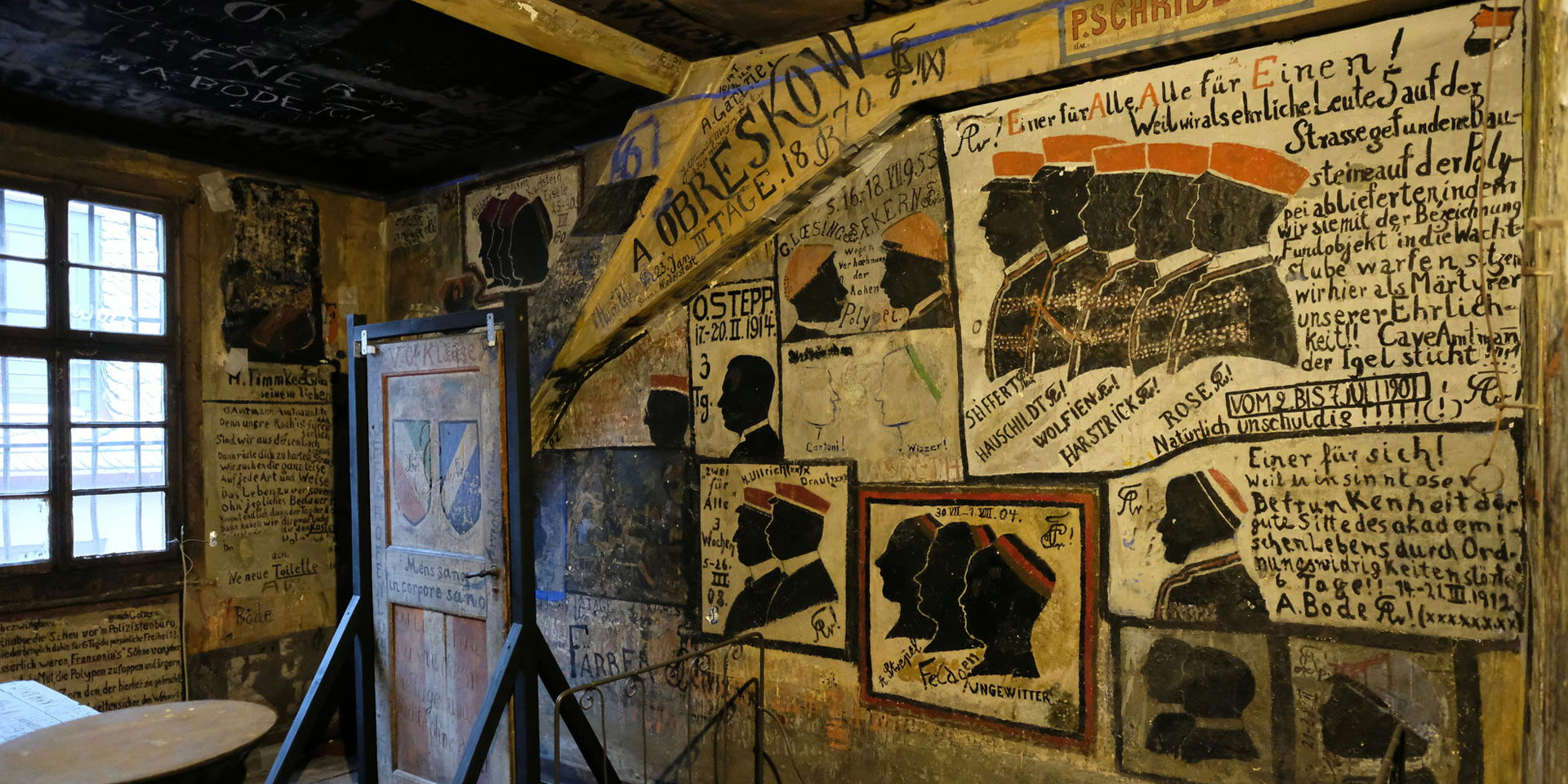 Ein historischer Raum mit Zeichnungen und Malereien an den Wänden. Mehrere Ausstellungsstücke, u. an. eine Tür in einem Gestell.