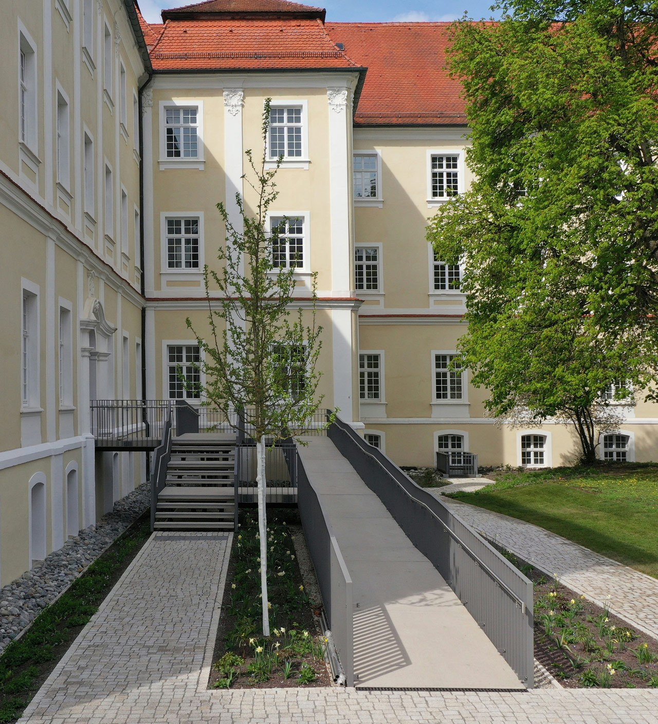 Ein Klostergebäude mit ockerfarbenen Wänden und weiß umrandeten Fenstern. Im Vordergrund eine lange Rampe.