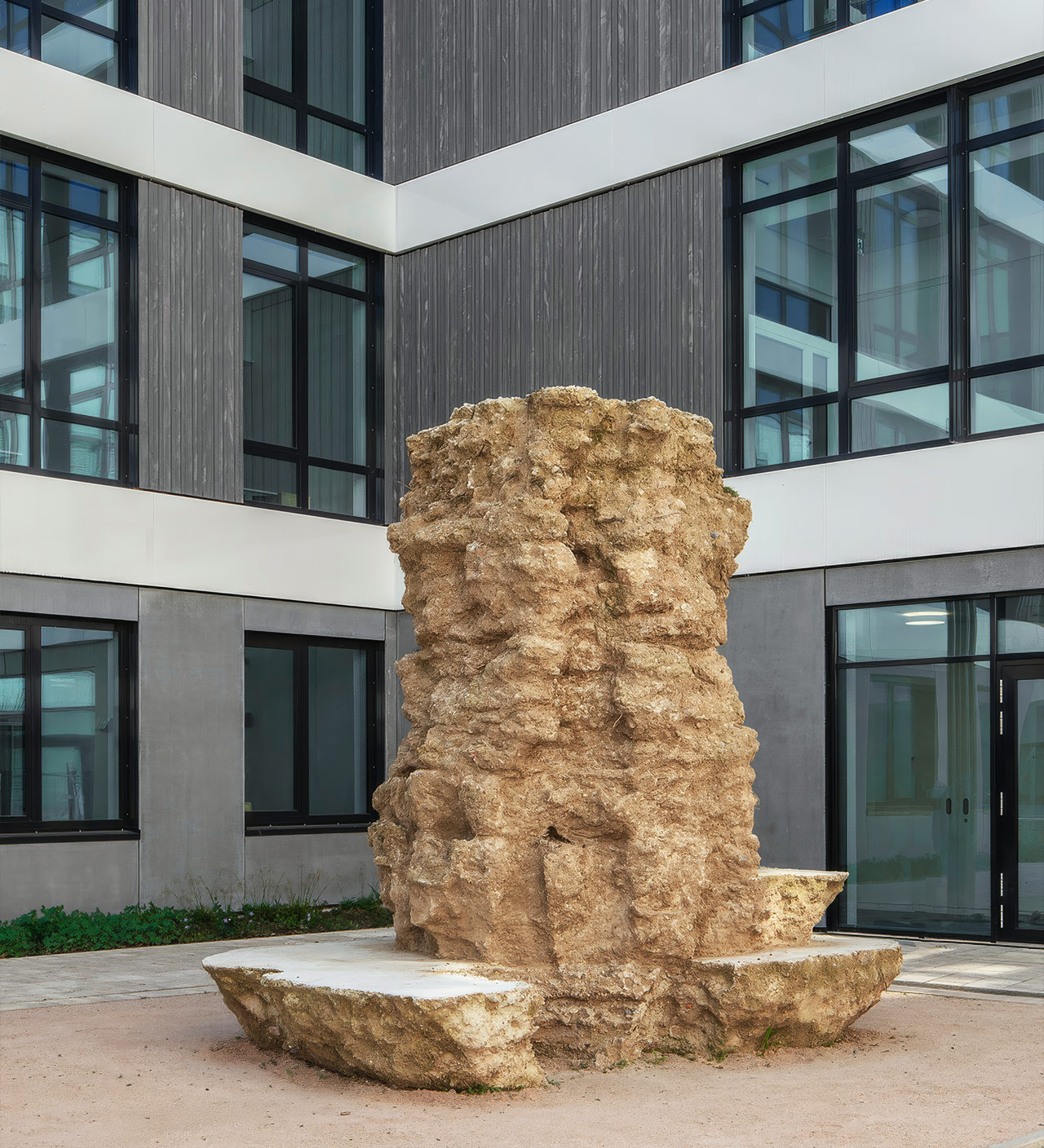 Skulptur aus Erd- und Betonmaterial im Hof des Neubaus, mit rauer Oberfläche und integrierten Sitzflächen. 