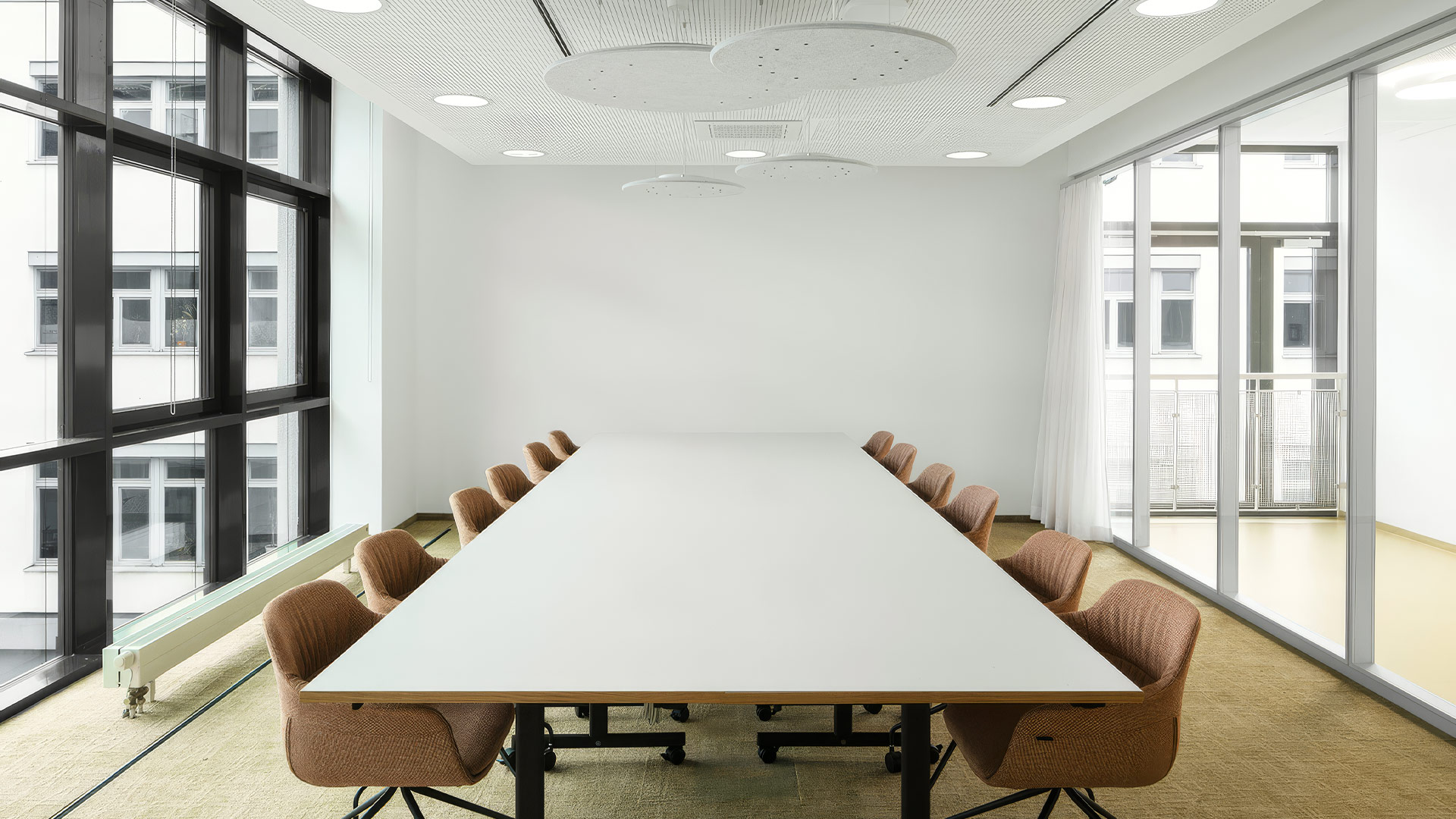 Heller Konferenzraum mit großem Tisch, umlaufenden Fenstern und modernen Deckenleuchten. 