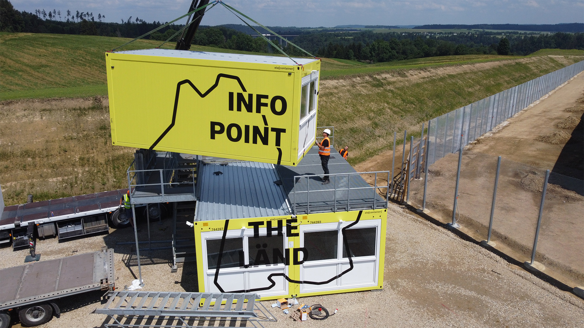 Drei gelbe Container mit Fenstern. Einer wird gerade auf zwei anderen positioniert. Darauf der Umriss von BaWü und Aufschriften: "Infopoint" und "THE LÄND"
