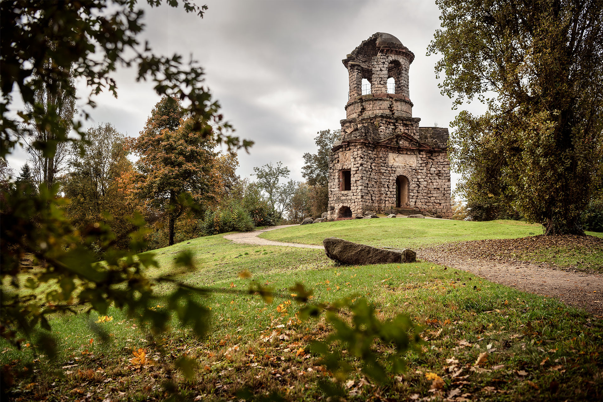 Ruine einer steinernen Kapelle umgeben von einem Park, düstere Stimmung, dunkle Wolken.
