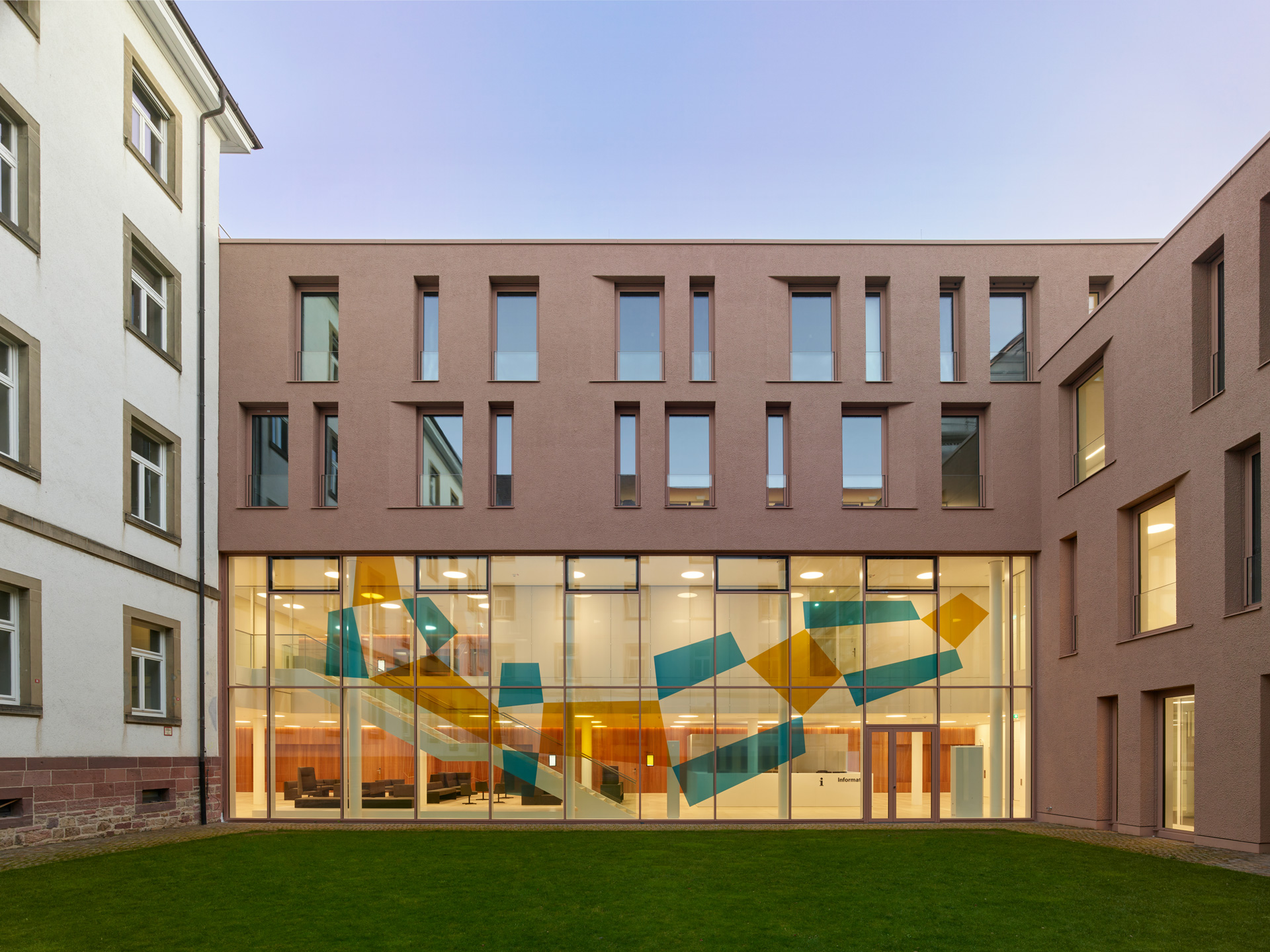 Ein an ein Bestandsgebäude angebauter Neubau im Dämmerlicht. Im Erdgeschoss eine große Fensterfläche mit bunten Farbfeldern.