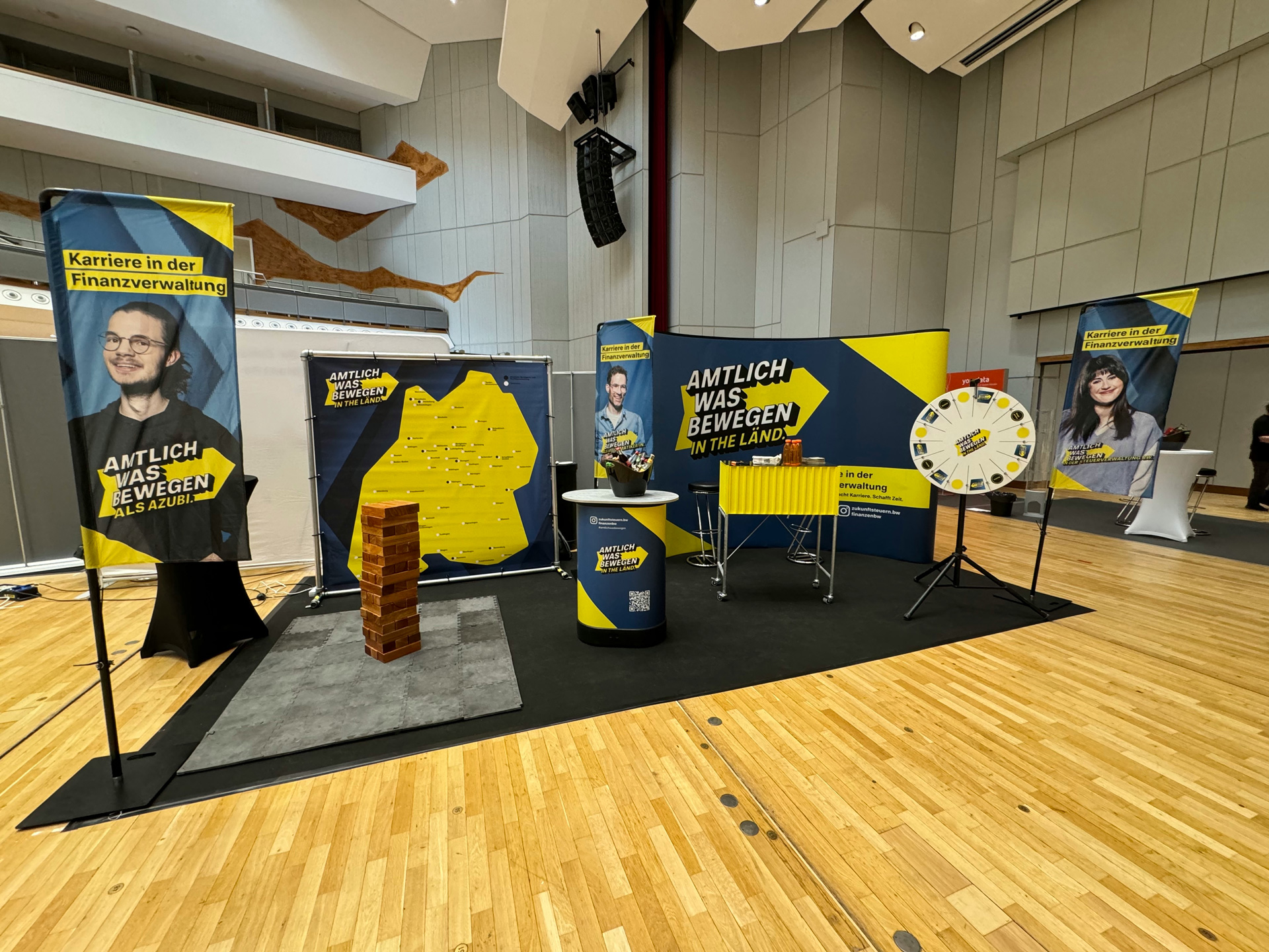 Eine Ausstellungsfläche auf Parkettboden, diverse Aufsteller mit der Aufschrift: Amtlich was bewegen, Stehtische, ein Glücksrad.
