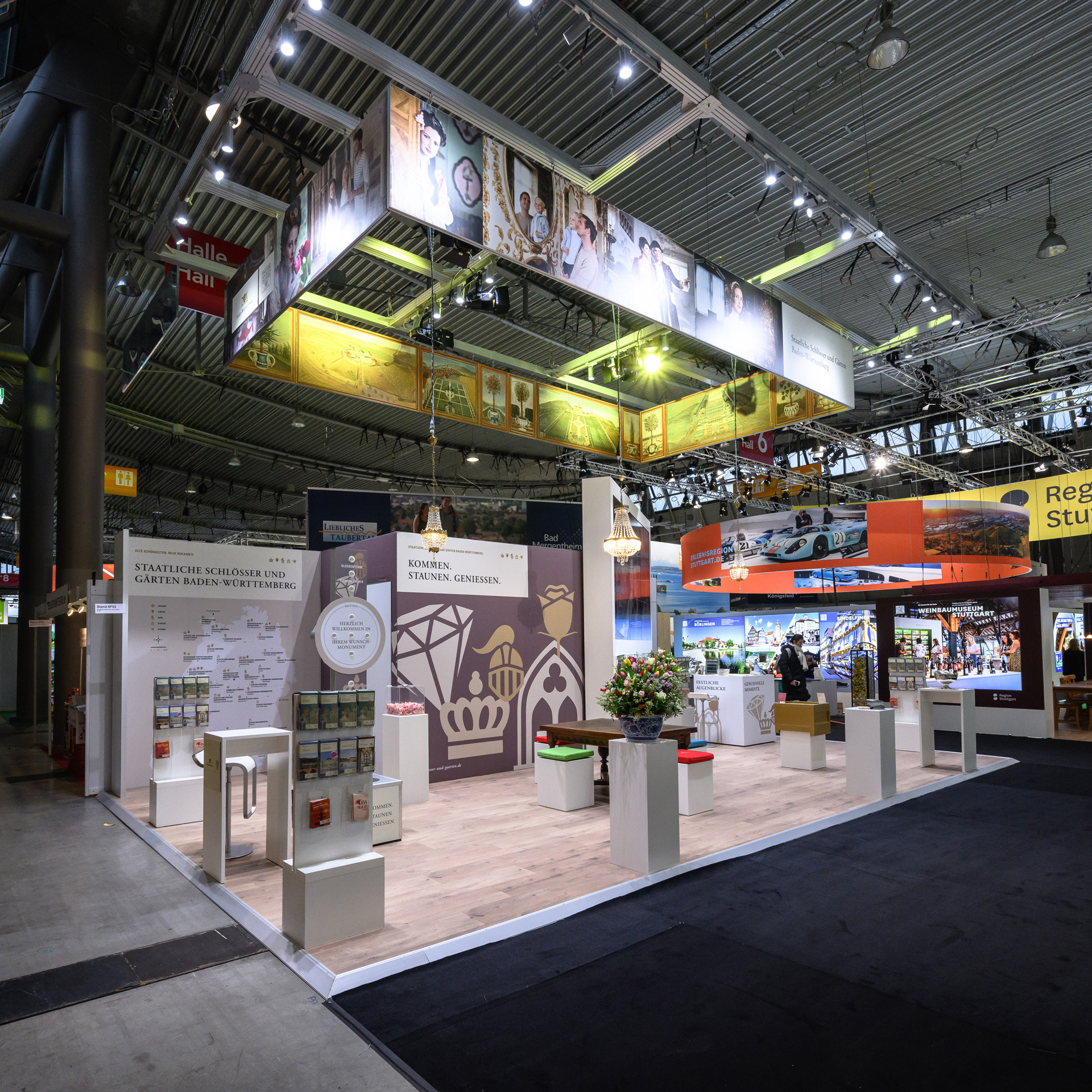 Eine Messehalle, ein ausgeleuchteter Messestand mit Sitzgelegenheiten, Stehtischen, Flyerständen und bebilderten Wänden.