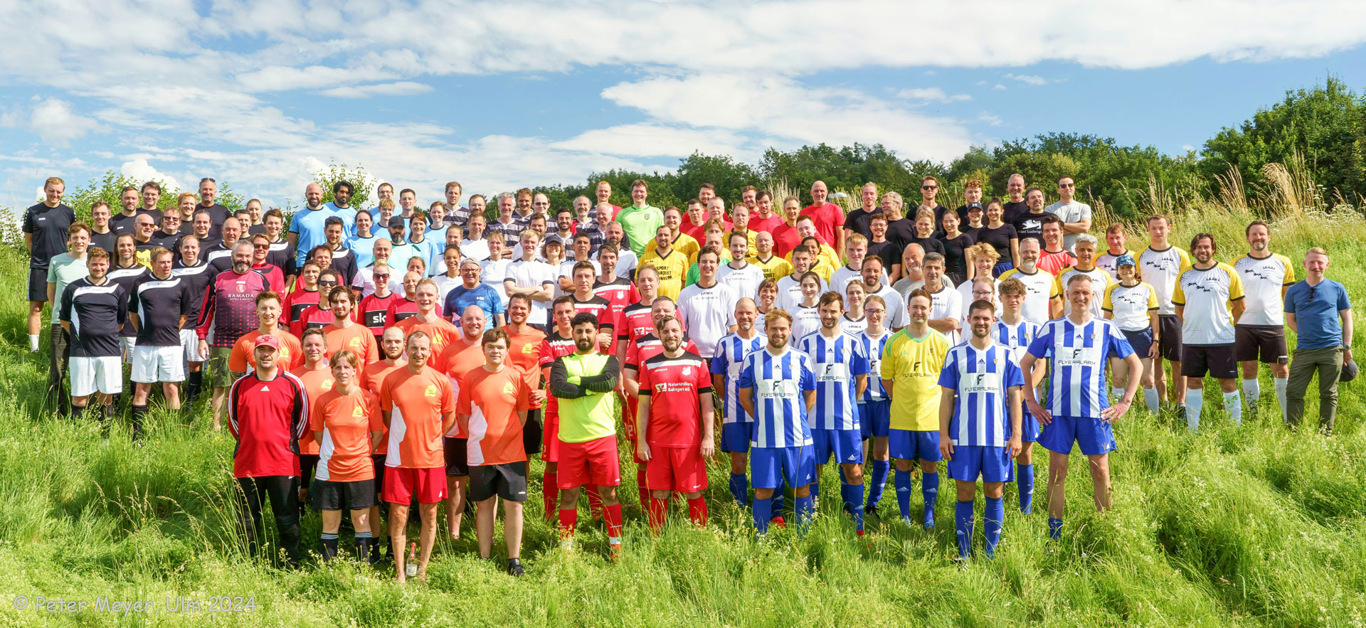 Mehrere Fußball-Teams im jeweils eigenen Mannschaftsdress stehen zum Gruppenbild auf einer hoch bewachsenen Wiese.