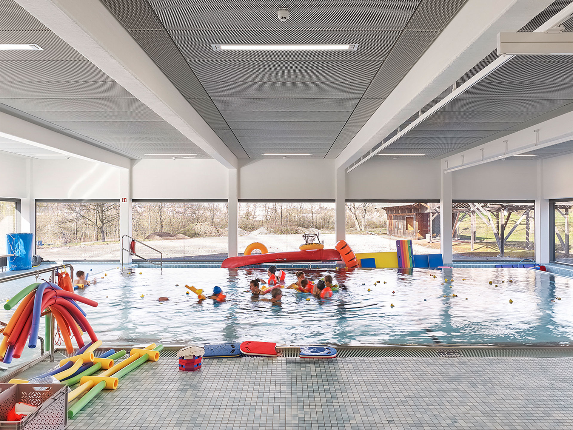 Ein Schwimmbad, im Becken Personen mit Schwimmflügeln, am Beckenrand bunte Pool-Nudeln und andere Schwimmutensilien.