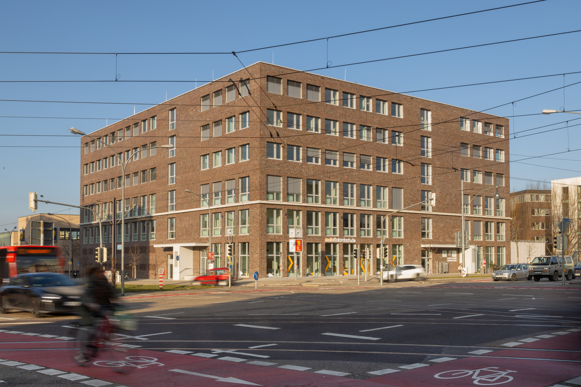 Blick auf ein 6-geschossiges Klinkerstein-Gebäude mit weißen Fensterrahmen und Flachdach über eine große Kreuzung hinweg.