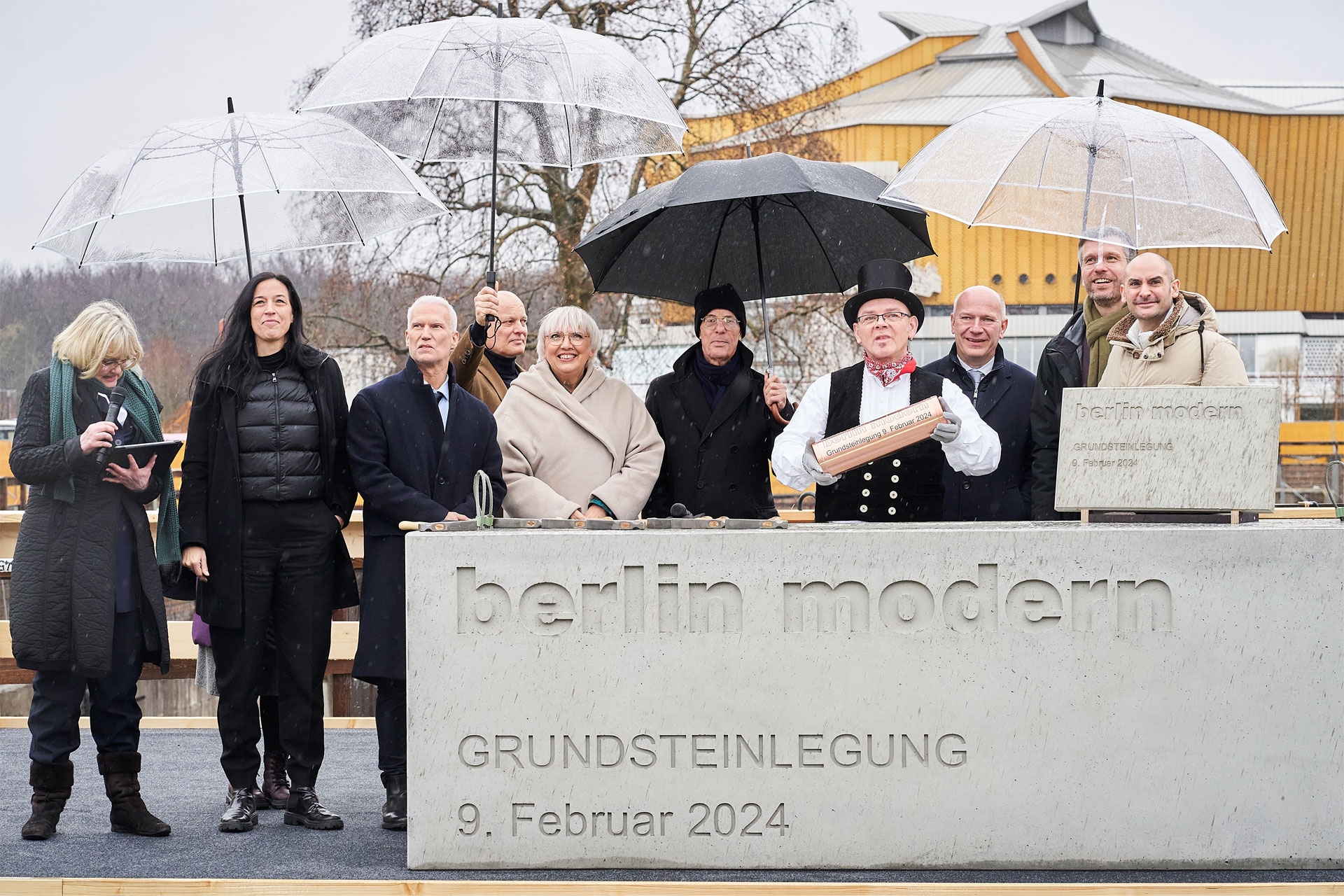 Ein Betonblock, Aufschrift berlin modern Grundsteinlegung 9.02.2024, dahinter eine Gruppe von Personen mit Regenschirmen.