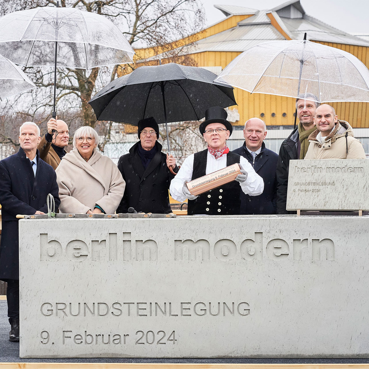Ein Betonblock, Aufschrift berlin modern Grundsteinlegung 9.02.2024, dahinter eine Gruppe von Personen mit Regenschirmen.