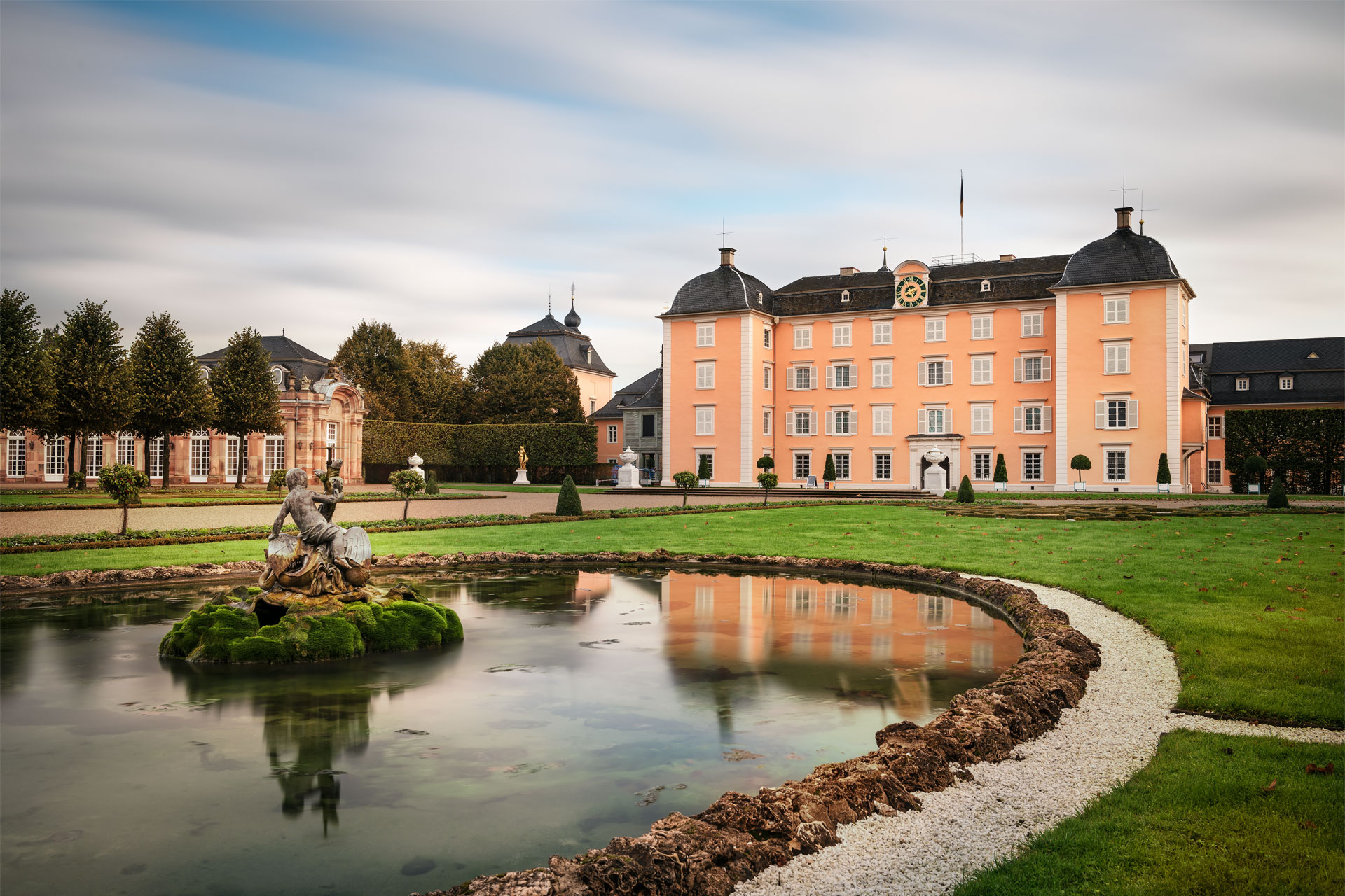 Ein Schloss mit ocker-farbener Fassade, davor ein Park mit Zierbecken, in dem sich das Schloss spiegelt.