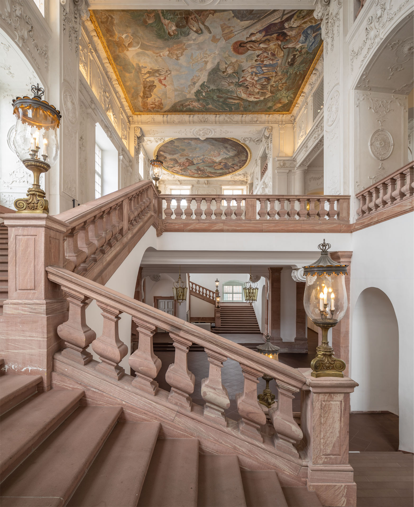Eine massive Treppe mit Marmor-Geländer in einem Schloss, prunkvolle Deckenmalerei und Stuck an den Wänden.