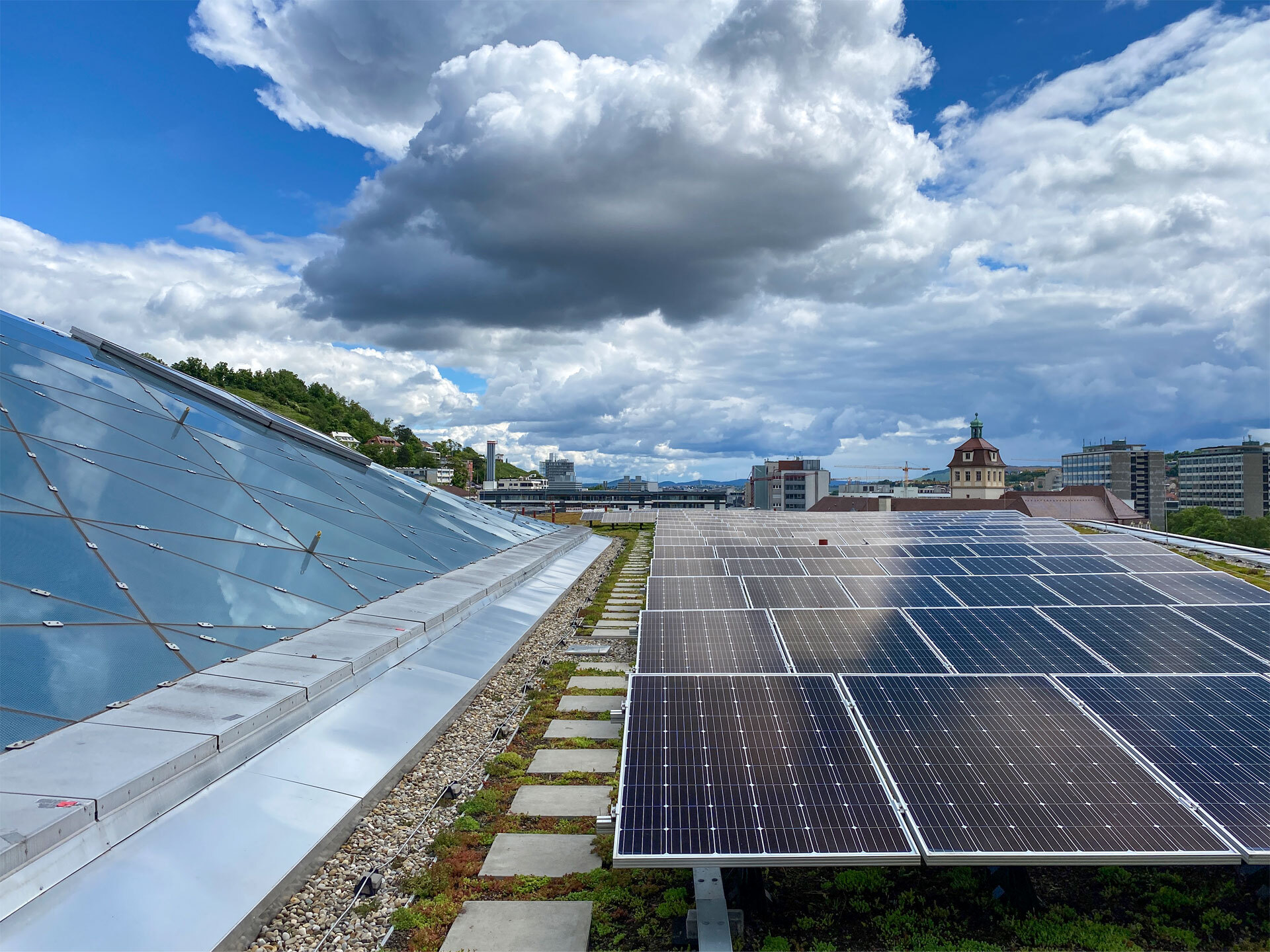Eine Dachfläche, rechts flach mit Photovoltaik-Modulen, links eine ansteigende Fläche aus Glaselementen, Wolken am Himmel.