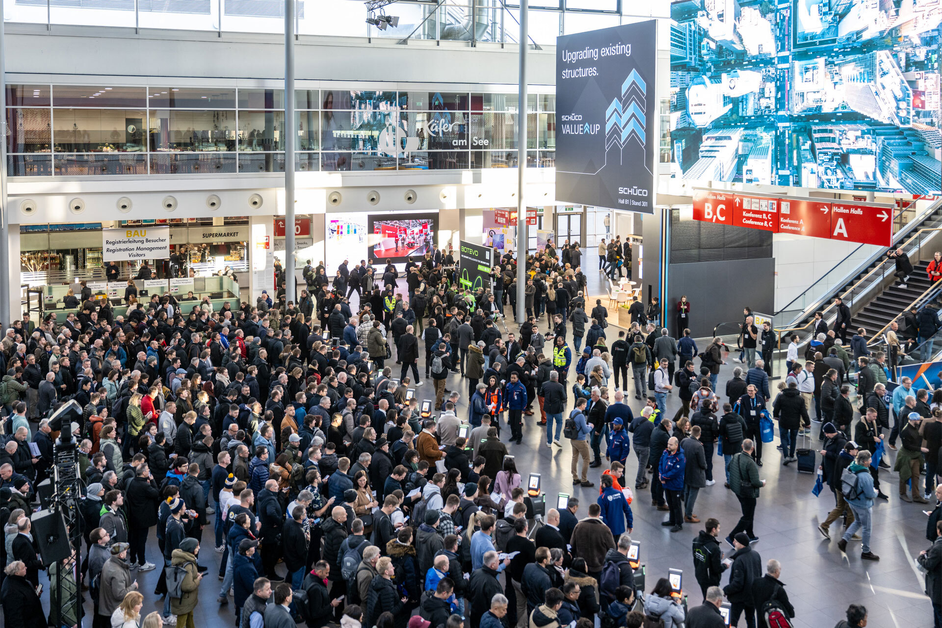 Eine Eingangshalle einer Messe mit vielen Menschen, rechts ein Treppenaufgang mit Wegweisern.
