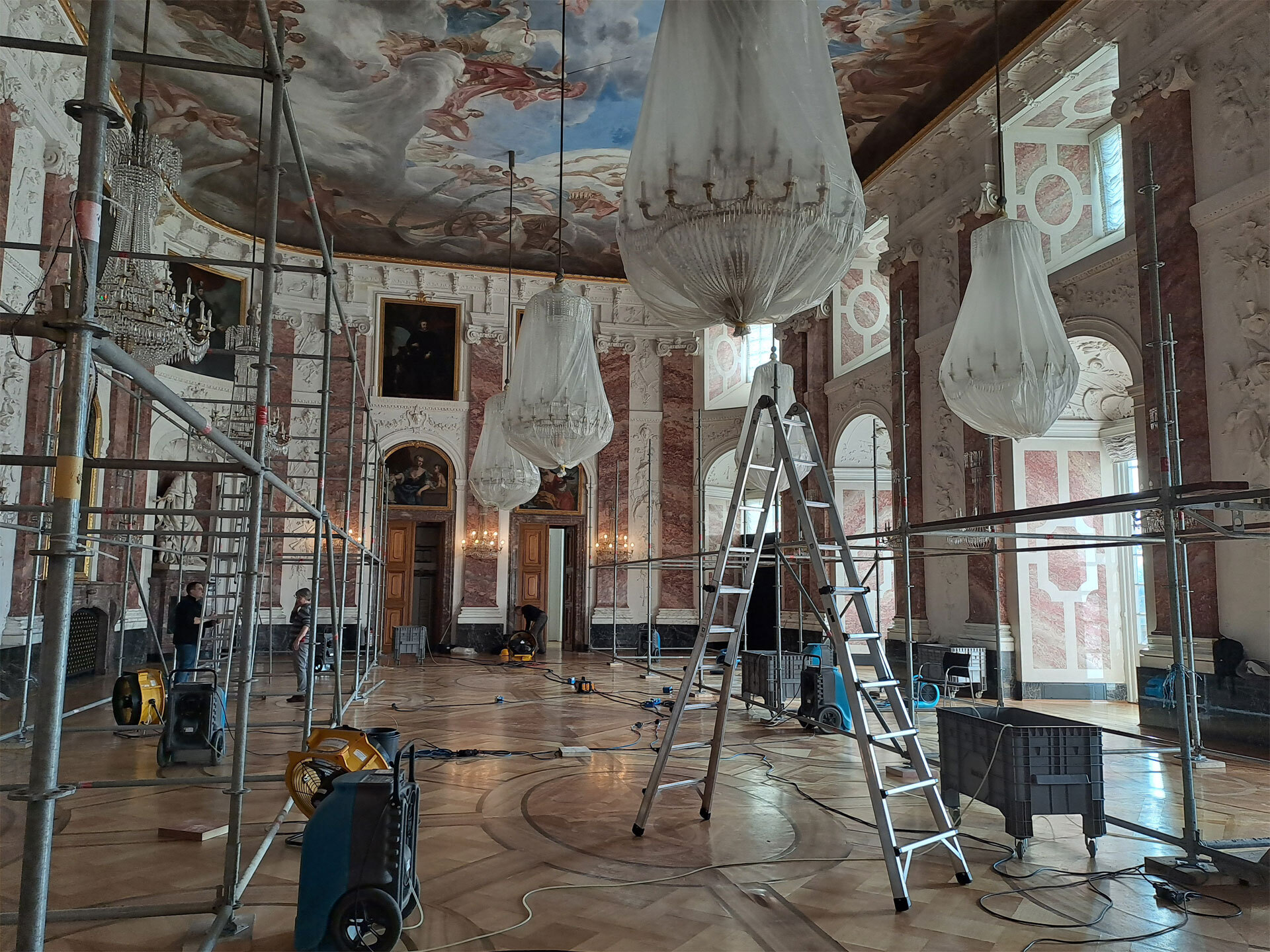 Ein prunkvoller Saal mit umhüllten Kronleuchtern, darin Gerüste, Leitern, Geräte und Stromkabel.
