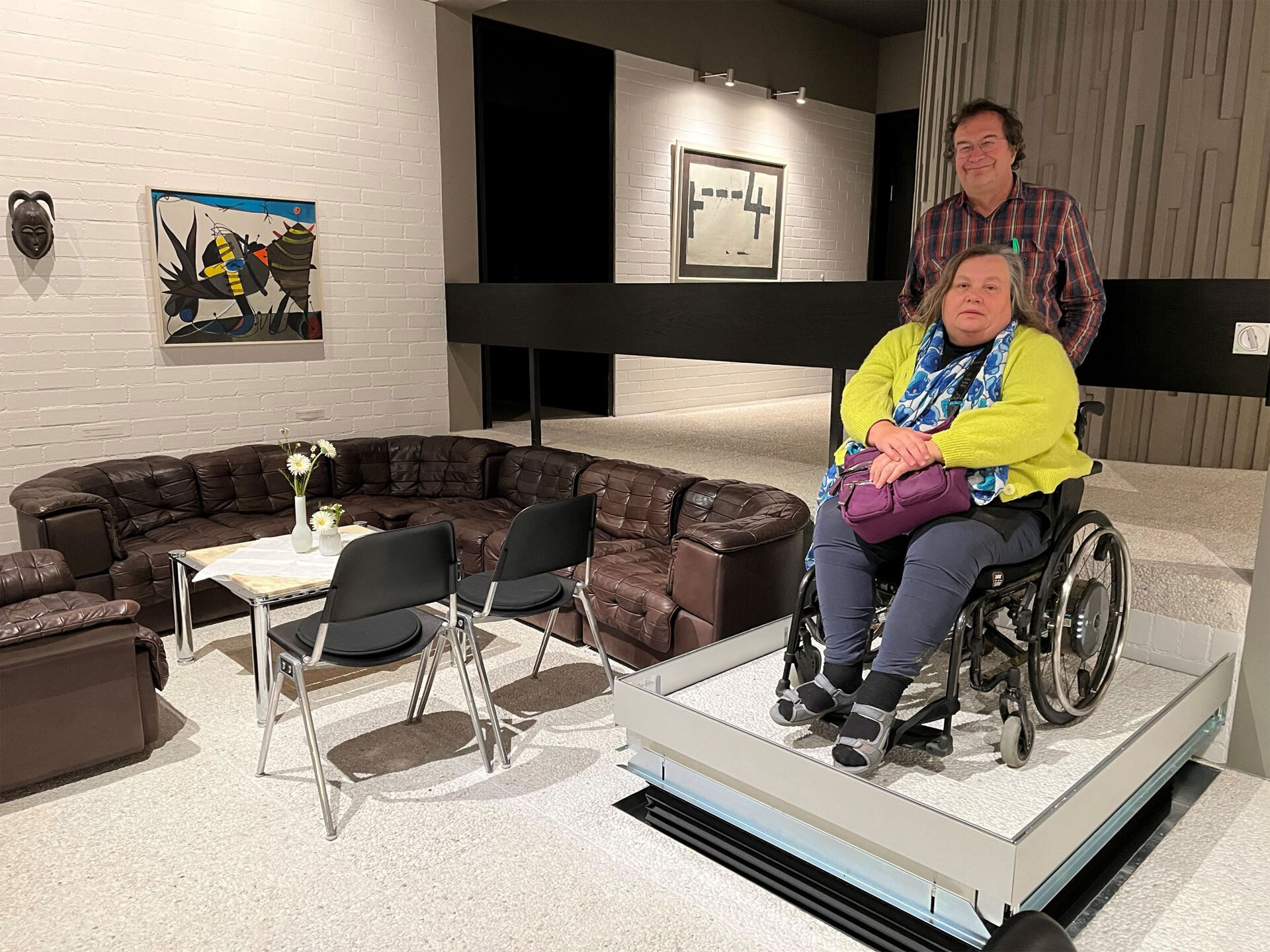 Ein Plattformlift transportiert zwei Personen, eine davon im Rollstuhl zwischen zwei Ebenen im Raum, daneben eine Sitzgruppe.