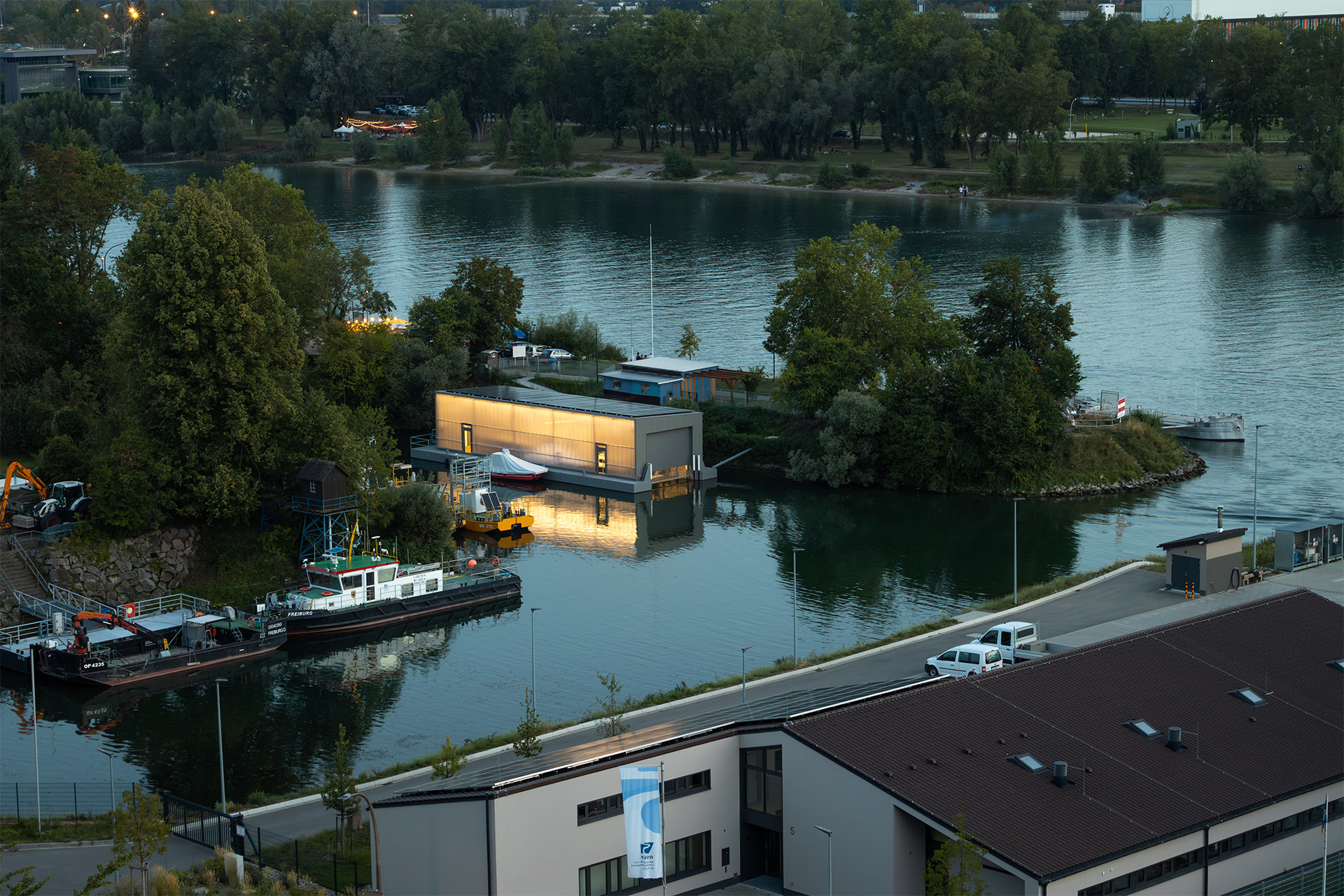 Ein Fluss mit Hafenbecken und Kaianlagen in der Dämmerung, die Umgebung trotz vieler Bäume technisch-industriell geprägt.