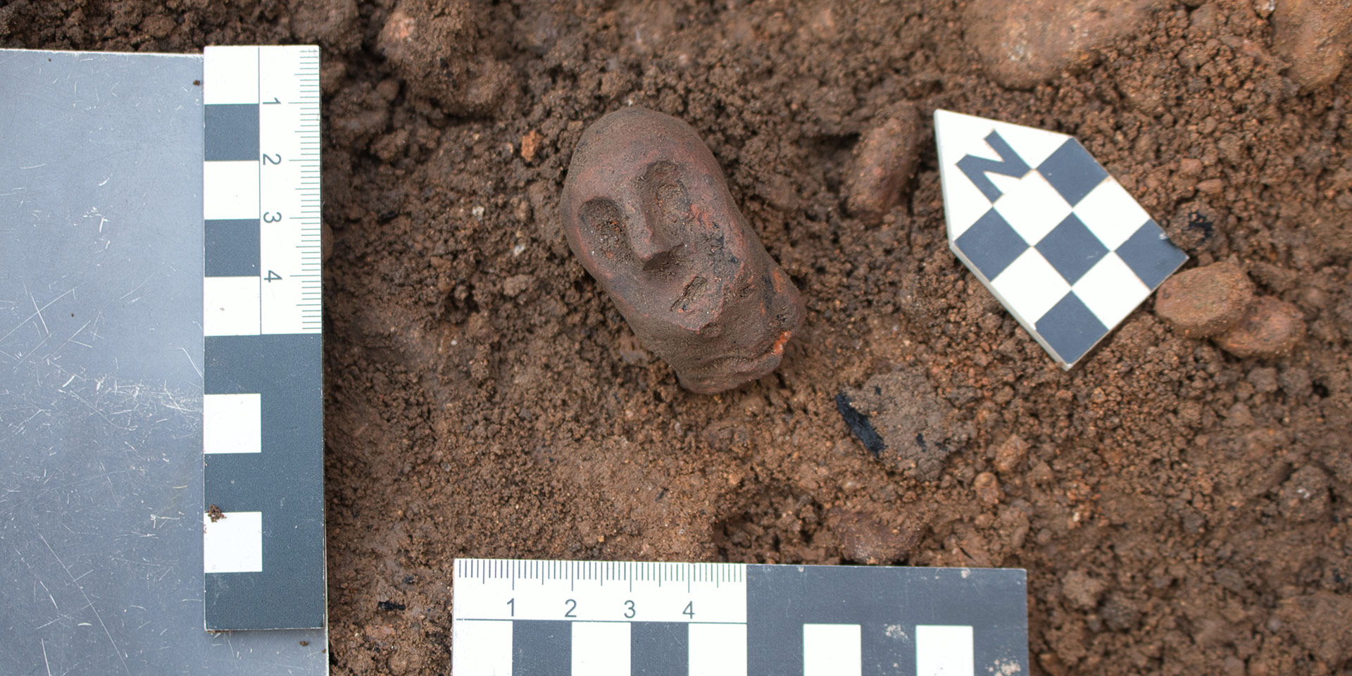 Ausgrabungsfund: roter Keramikkopf einer mittelalterlichen Figur mit Gugel, 4,8 cm groß, in der Ausgrabungsschicht mit Maßstäben und Nordpfeil.