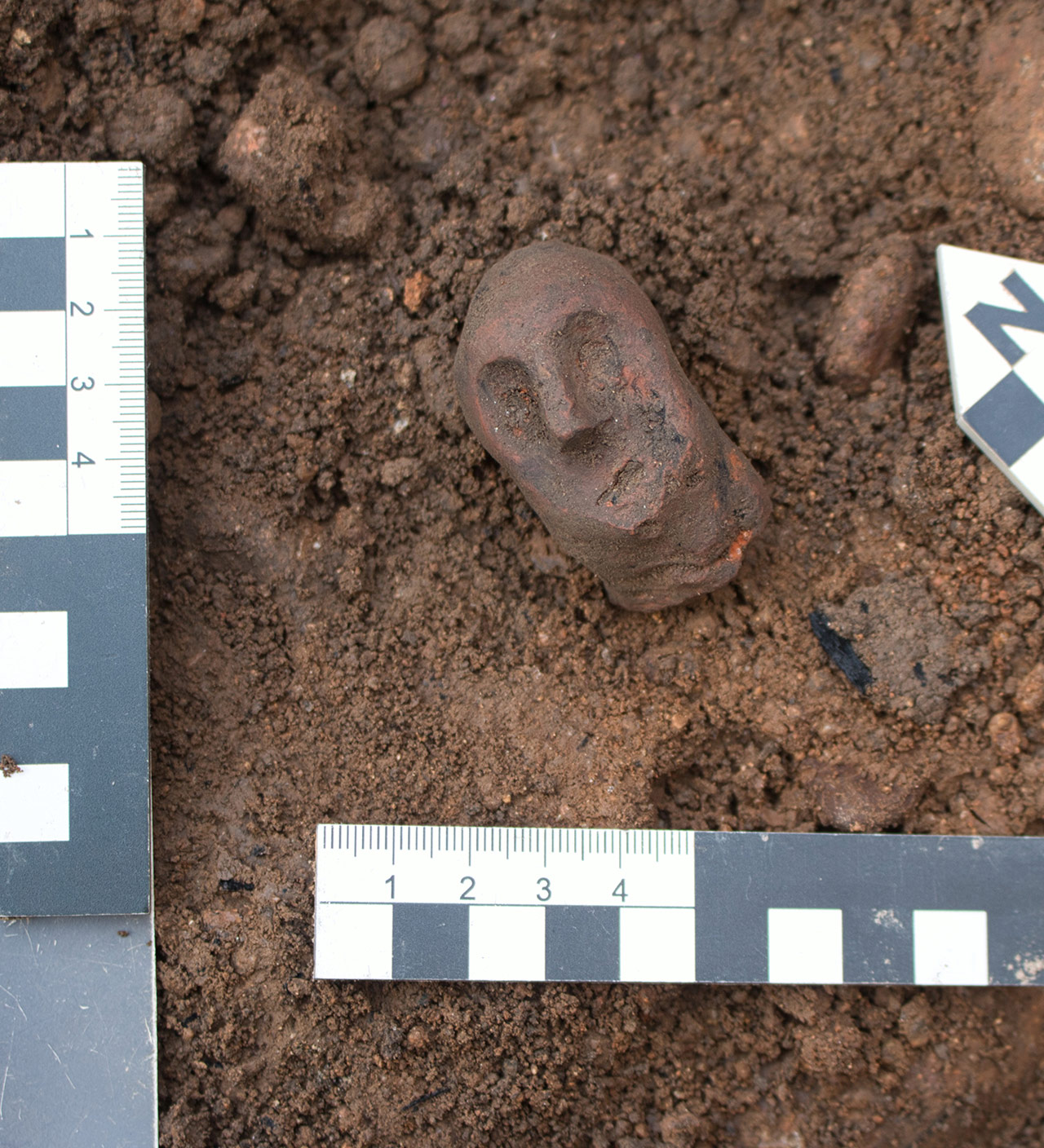Ausgrabungsfund: roter Keramikkopf einer mittelalterlichen Figur mit Gugel, 4,8 cm groß, in der Ausgrabungsschicht mit Maßstäben und Nordpfeil.