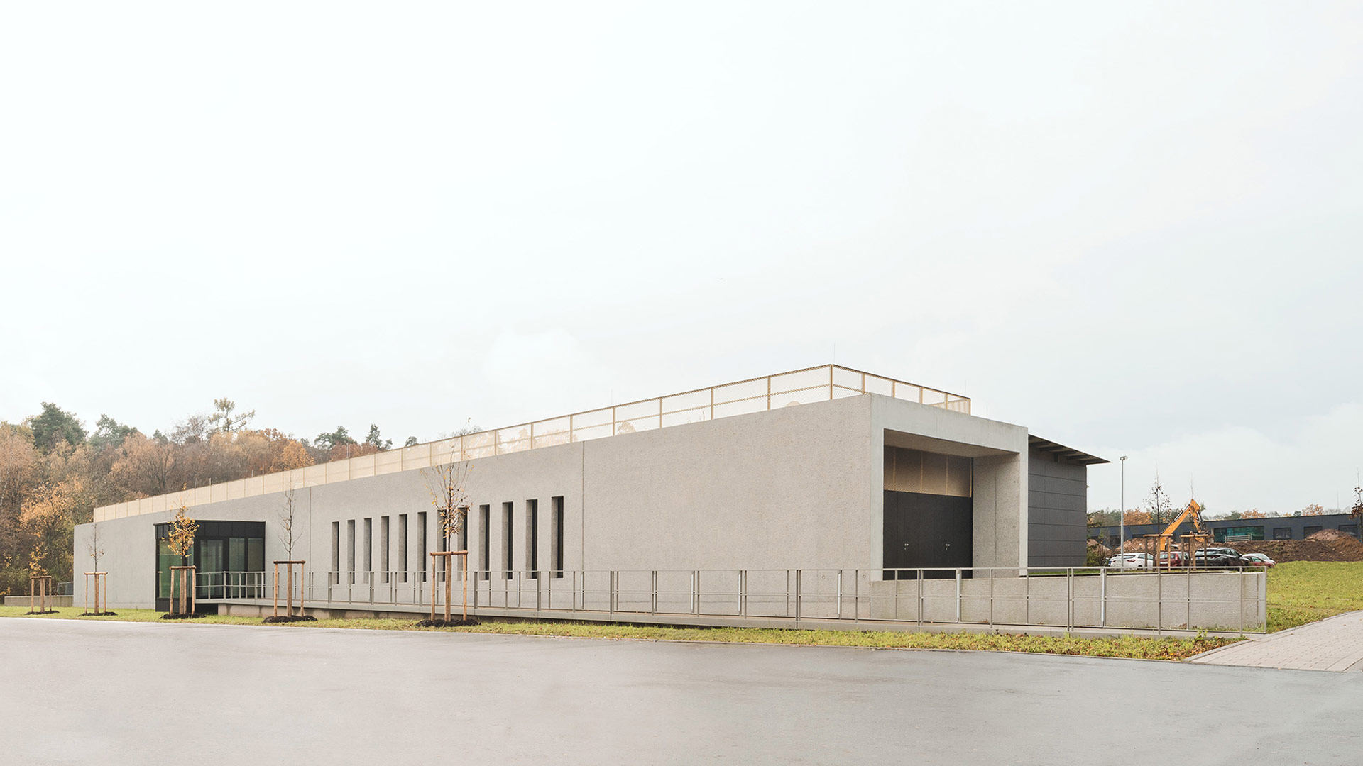 Ein langes Gebäude mit Sichtbeton-Fassade, auf dem Flachdach: ein Geländer. Davor eine Asphaltfläche.