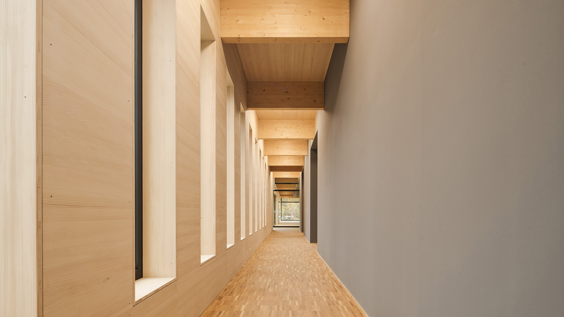 Ein Flur mit Parkettboden, rechts eine grau-braune, links eine holzvertäfelte Wand mit Fenstern, die Decke aus Holz. Warme Farben.