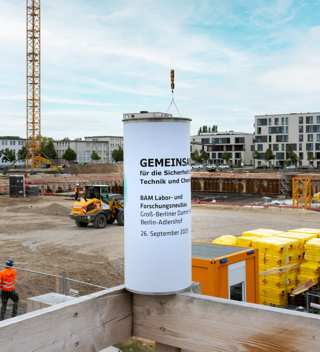 Im Hintergrund eine Baustelle mit Turmdrehkran. Im Vordergrund steht auf einem Holzgeländer eine Zeitkapsel.