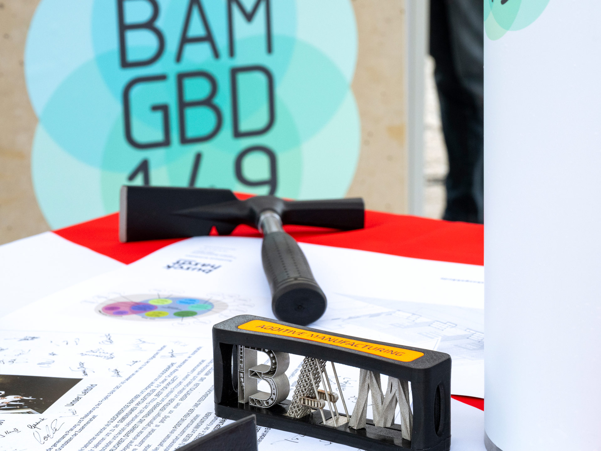 Ein Tisch mit u.a. einem Maurerhammer, einigen Dokumenten, einem 3D-gedruckten Gegenstand, der die drei Buchstaben BAM zeigt.