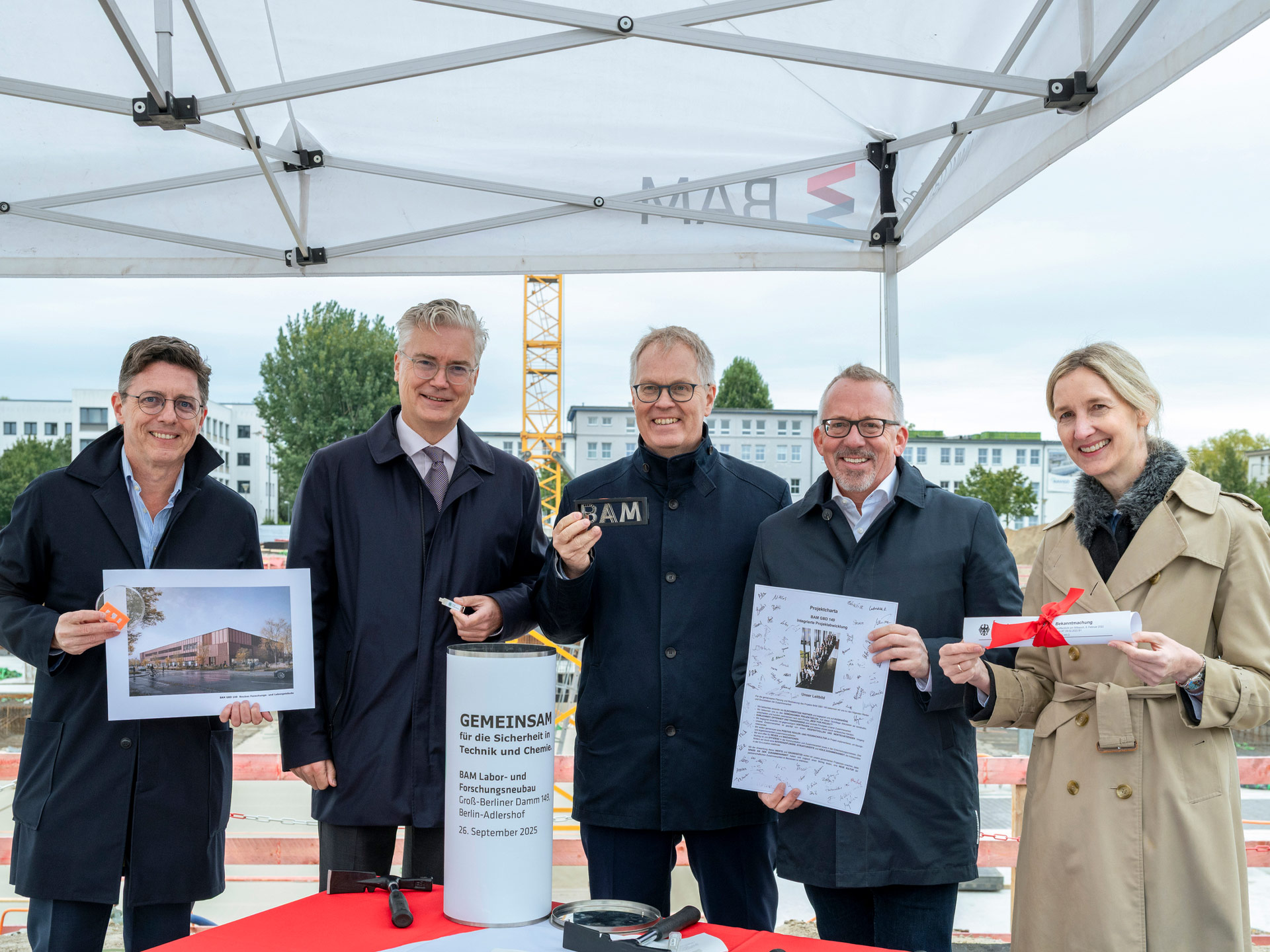 Gruppenbild von fünf Personen, sie halten Gegenstände und Dokumente in die Kamera, im Hintergrund eine Baustelle.