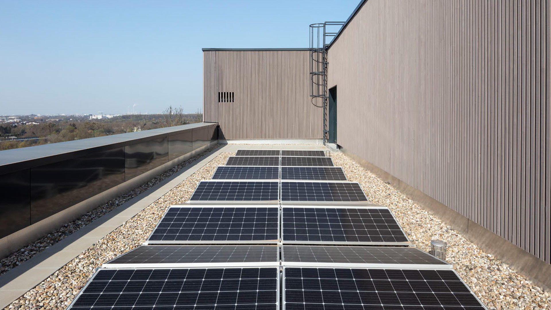 Detailaufnahme der Photovoltaik-Anlage auf dem Dach des Laborgebäudes. Reihen von Solarmodulen auf Kiesbett vor der Fassade des Neubaus.