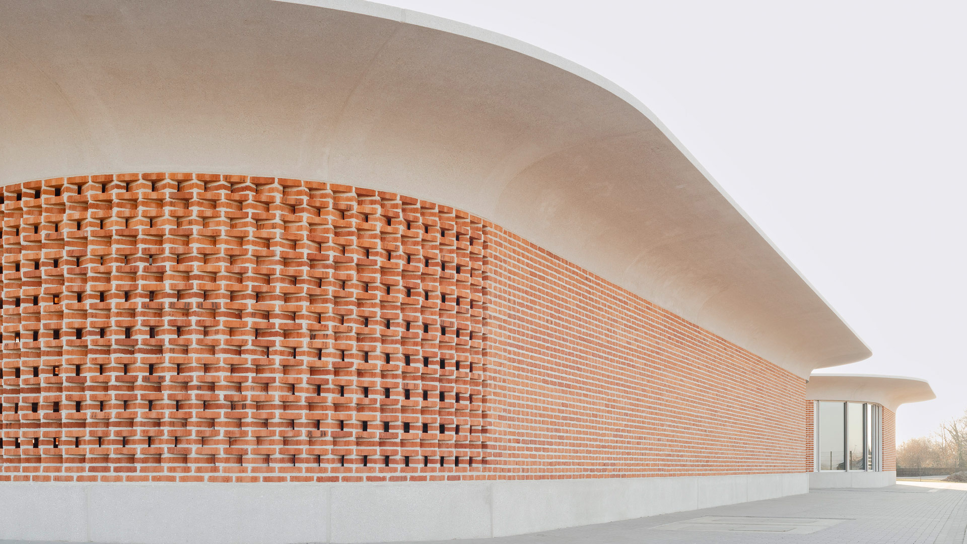 Zwei ovale Gebäude mit auskragendem Betondach und abgesetzter Klinkermauer, eins fensterlos, das andere mit bodentiefen Fenstern.