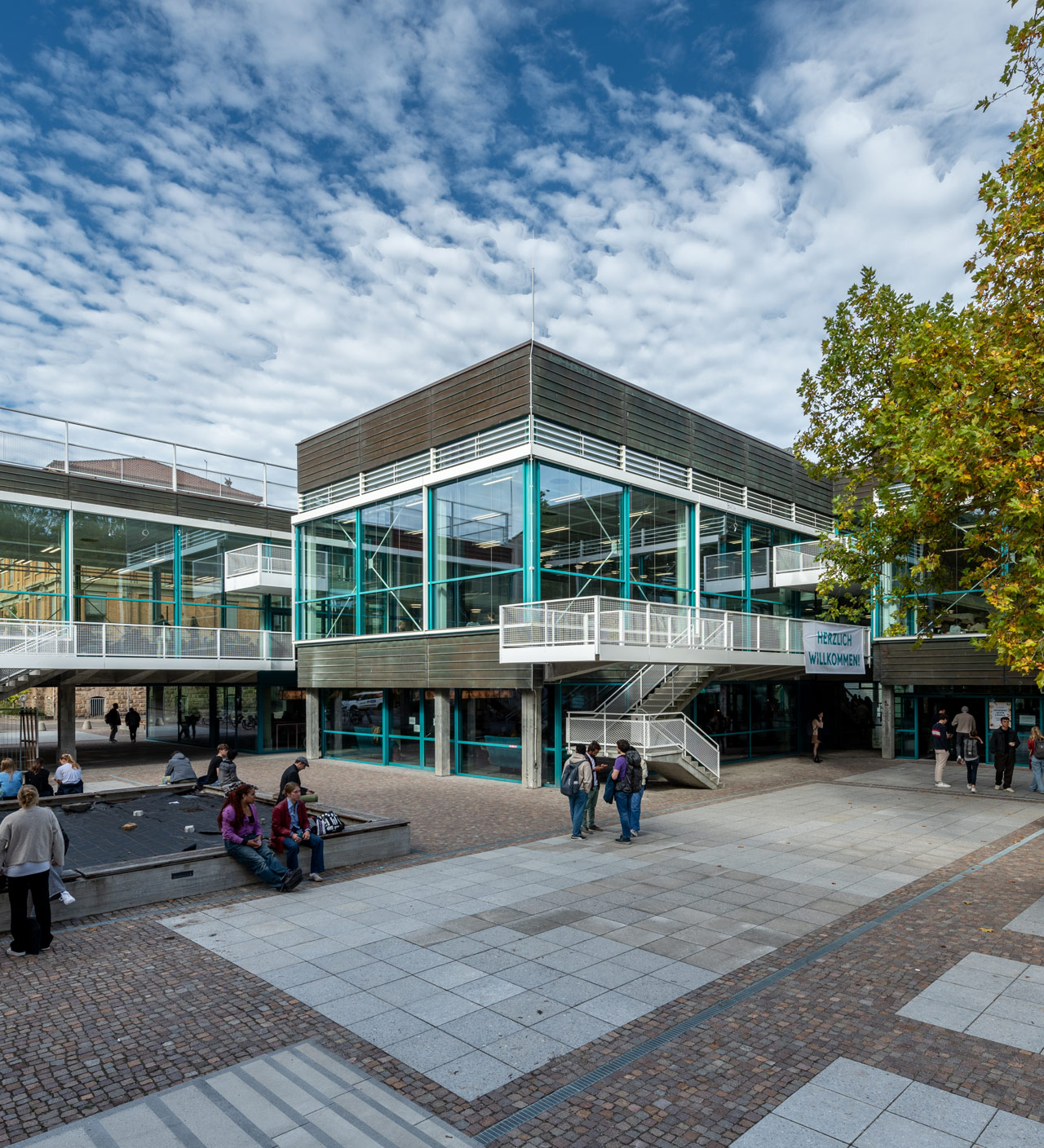Im Hof, vor einem 2-stöckigen Flachdachgebäude mit türkis-farbenen Fenstern sitzen, stehen und laufen verschiedene Personengruppen.
