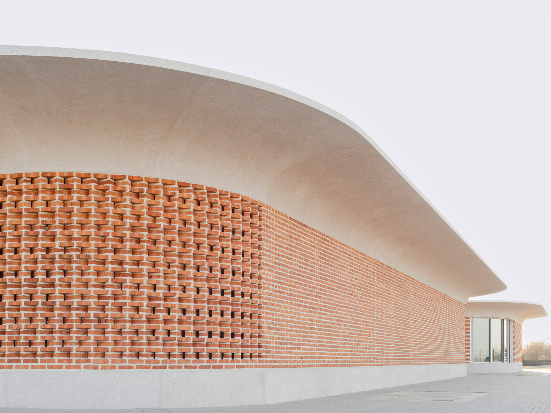 Zwei ovale Gebäude mit auskragendem Betondach und abgesetzter Klinkermauer, eins fensterlos, das andere mit bodentiefen Fenstern.