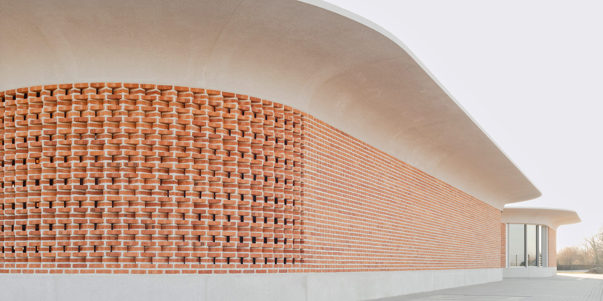 Zwei ovale Gebäude mit auskragendem Betondach und abgesetzter Klinkermauer, eins fensterlos, das andere mit bodentiefen Fenstern.