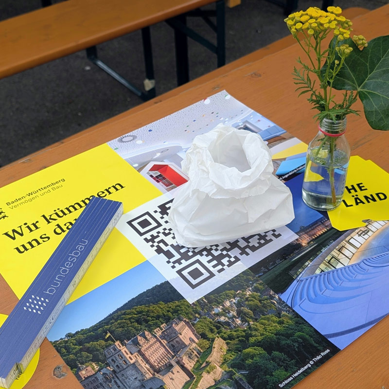 Tischdekoration einer VBV Jubiläumsfeier mit Infomaterialien, QR Code und kleiner Vase als Teil des sommerlichen Festambientes. 