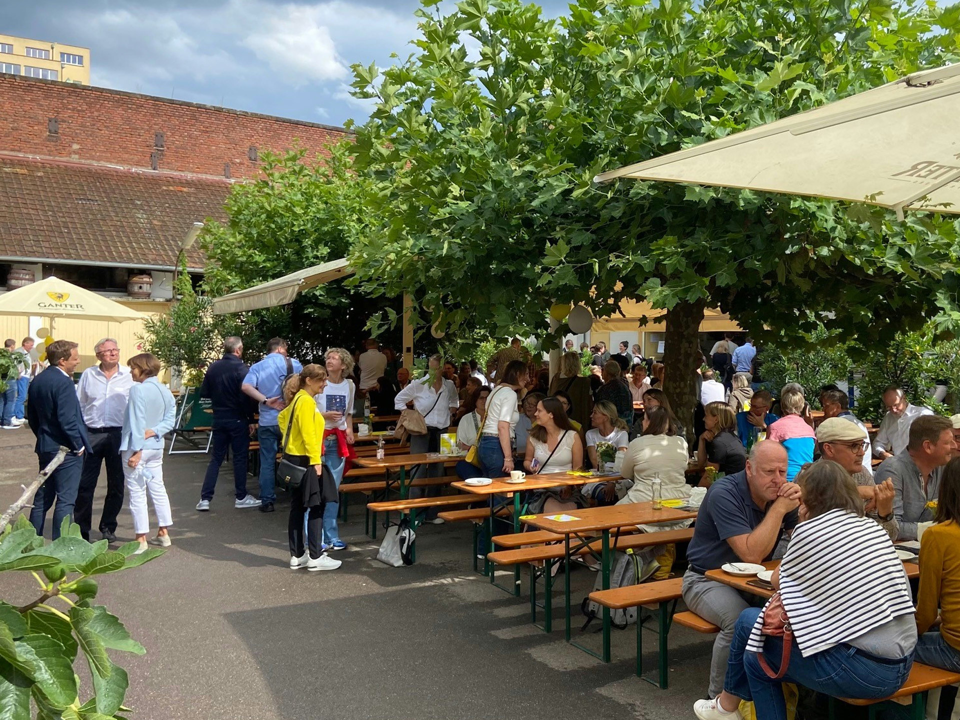 Viele Menschen sitzen in einem schattigen Außenbereich an langen Holztischen, essen und unterhalten sich in entspannter Atmosphäre unter Bäumen und Sonnenschirmen. 