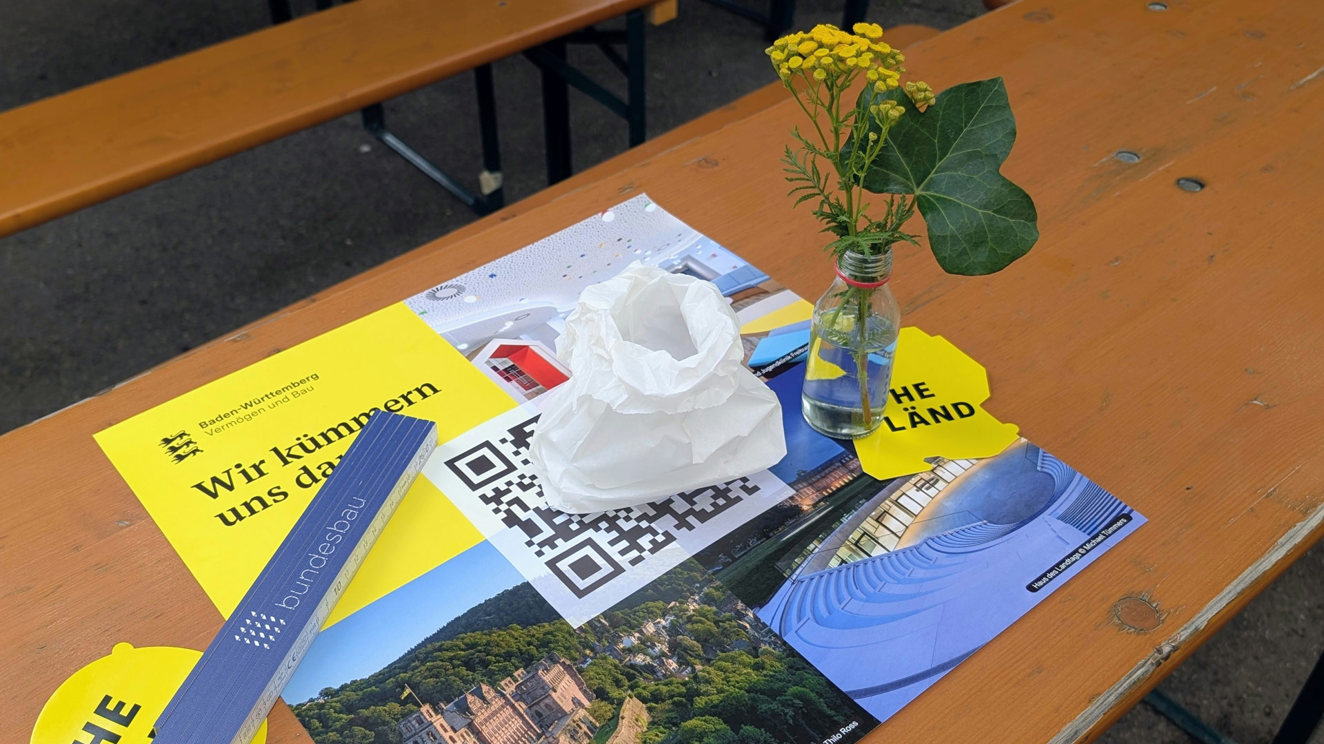 Tischdekoration einer VBV Jubiläumsfeier mit Infomaterialien, QR Code und kleiner Vase als Teil des sommerlichen Festambientes. 