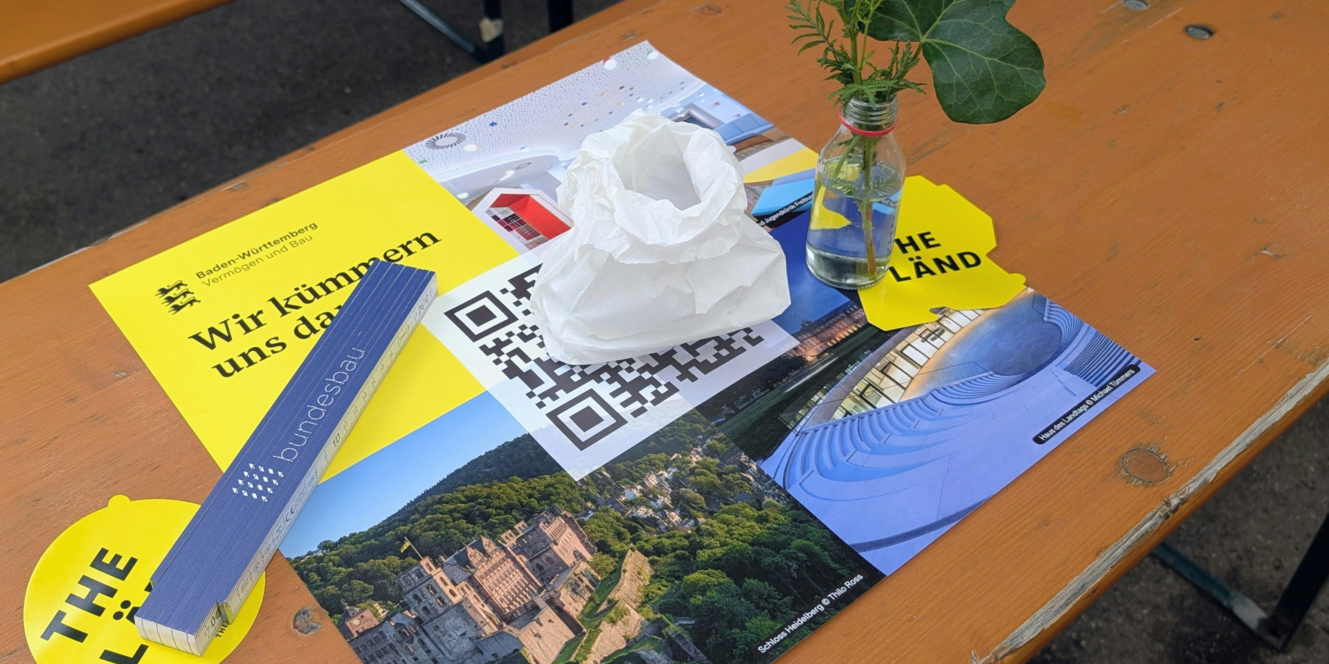 Tischdekoration einer VBV Jubiläumsfeier mit Infomaterialien, QR Code und kleiner Vase als Teil des sommerlichen Festambientes. 