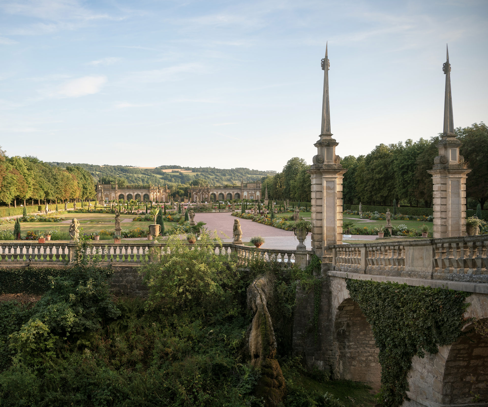 Im Vordergrund eine steinerne Brücke und ein wild bewachsender Hang. Dahinter der vielfältig angelegte Schlossgarten Weikersheim.