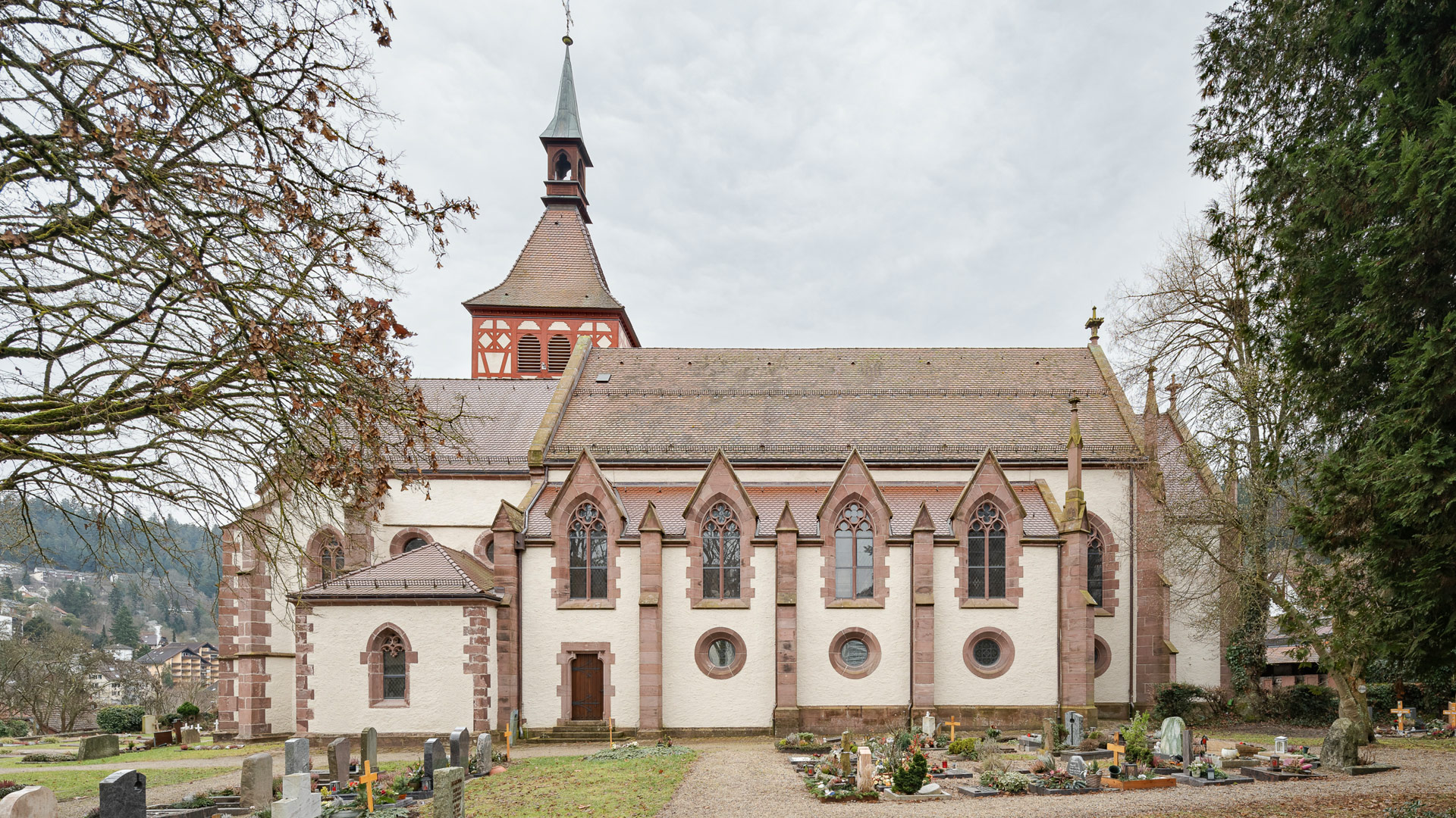 Weiß verputzter Kirchenbau mit rotem Sandstein, gotischen Fenstern und schlankem Dachreiter, vom Friedhof aus fotografiert. 