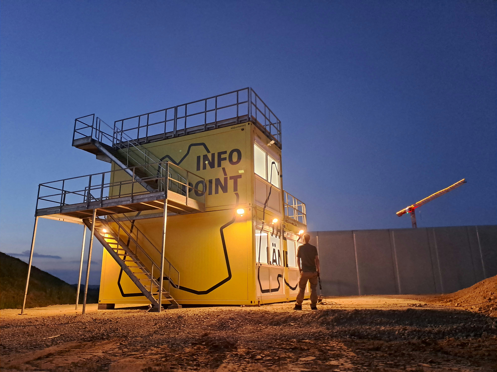 Eine Person steht in der Dämmerung vor drei gelben Containern mit einer Außentreppe, die auf das Dach führt. Aufschrift: "Infopoint" und "The Länd".
