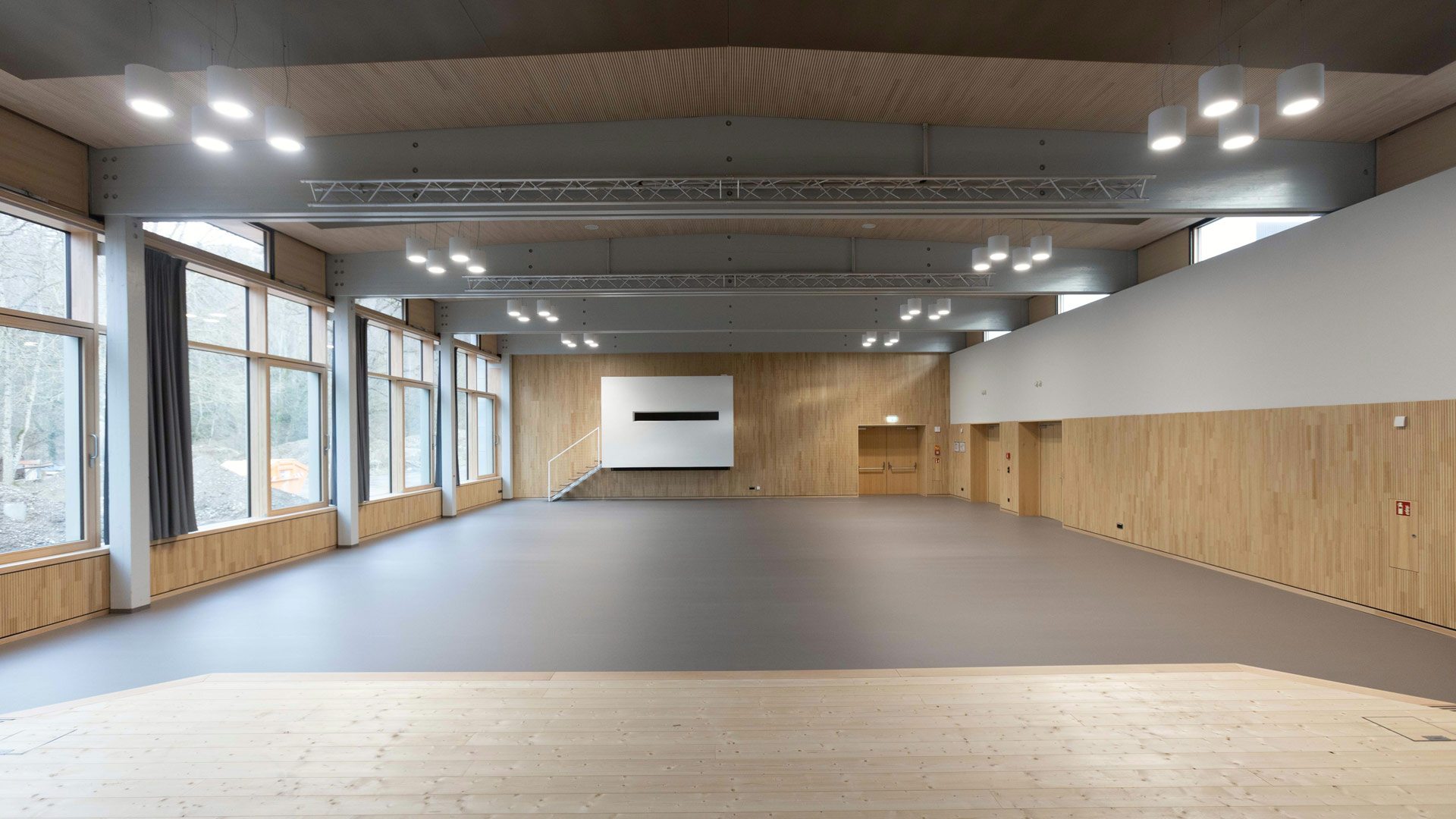 Eine Halle mit Holzboden, die Wände nur zum Teil aus Holz. Links eine Fensterfront mit grauen, gerafften Vorhängen.
