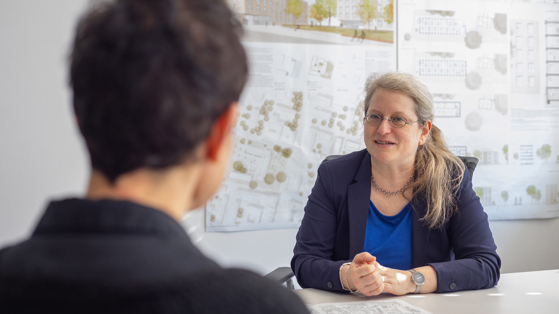 -	Projektleiterin Katharina Birkenmeier sitzt am Tisch und hört zu, im Hintergrund Baupläne. 
