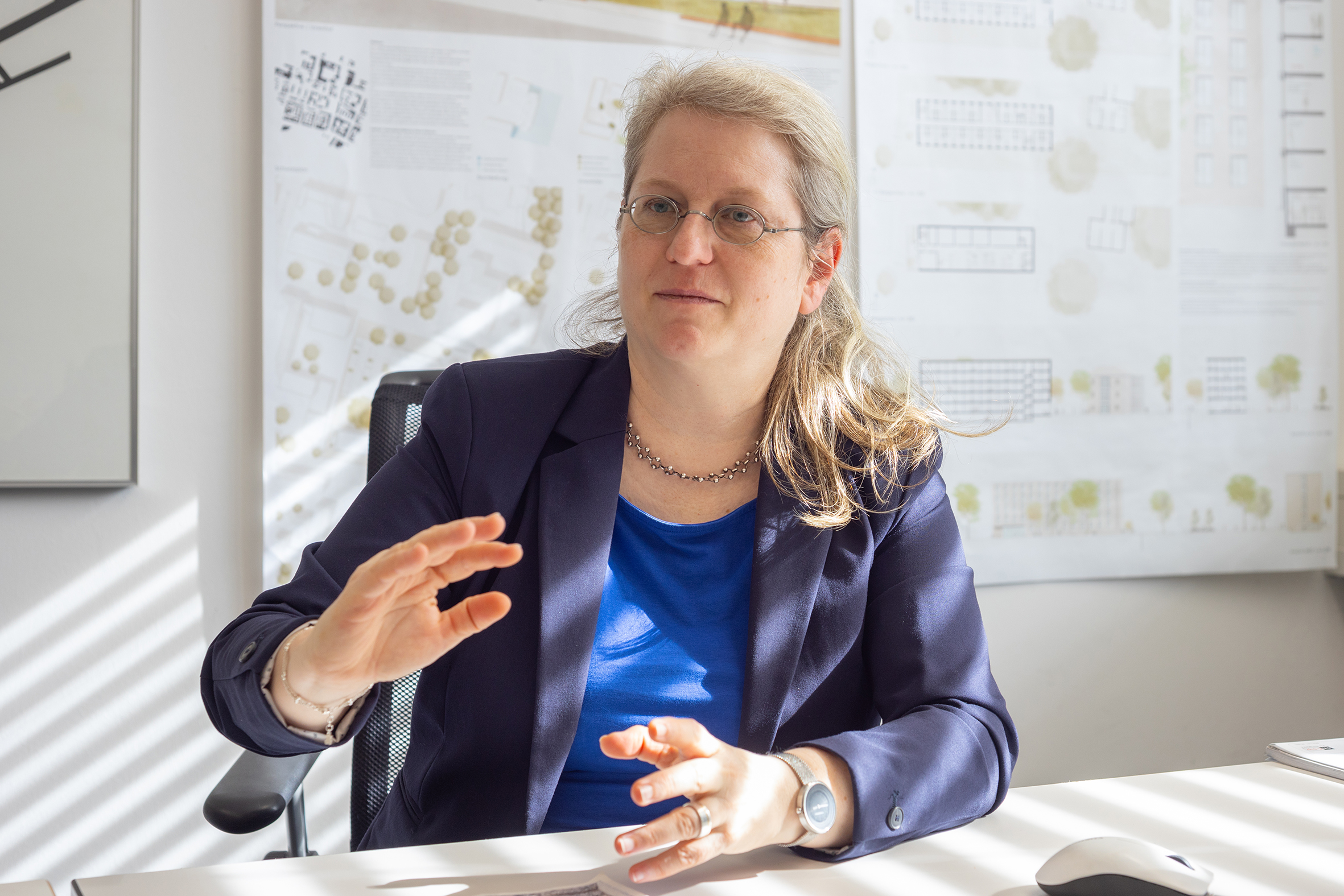-	Katharina Birkenmeier erklärt mit beiden Händen, im Hintergrund Baupläne.