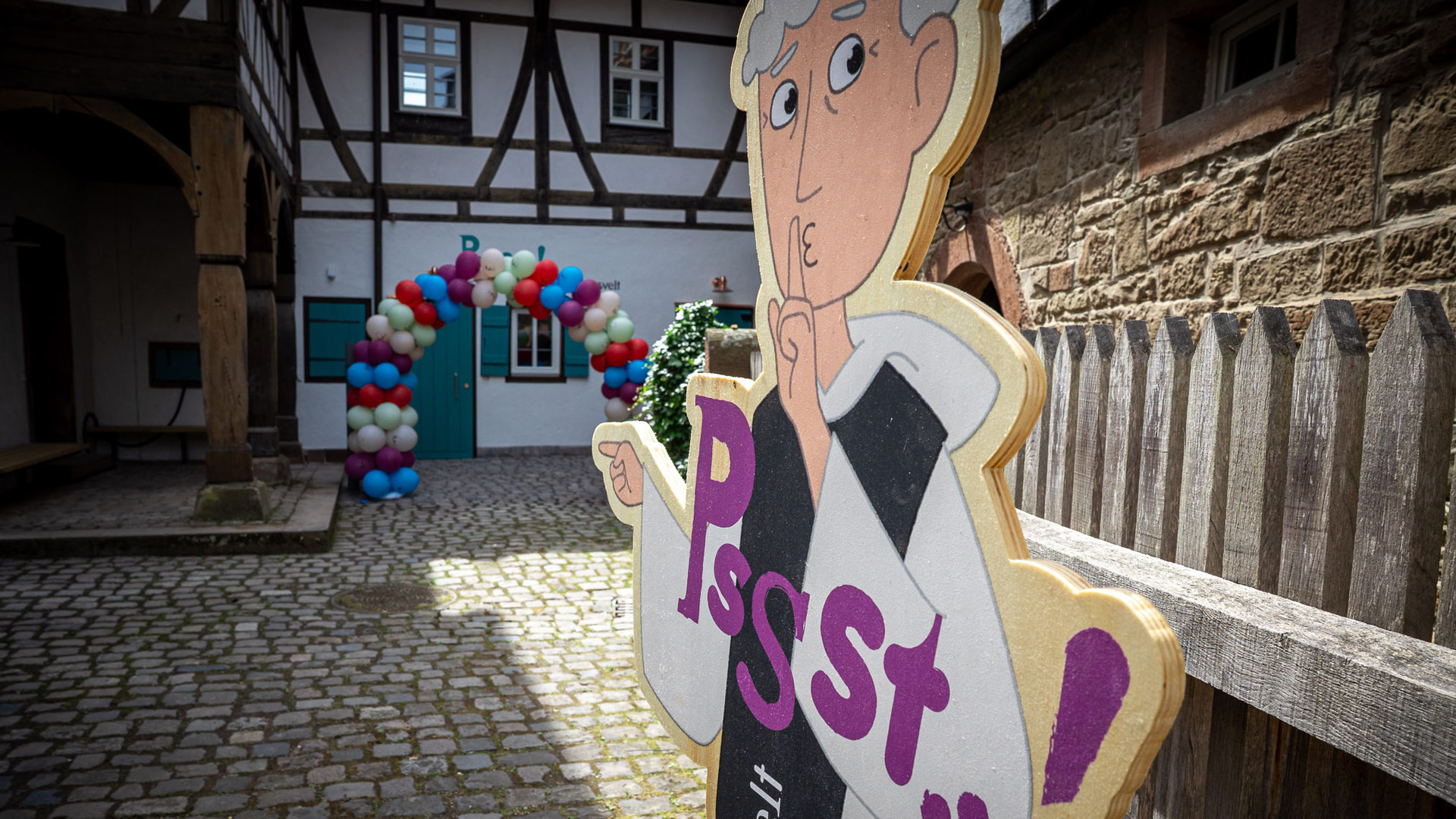 Im Vordergrund eines Hofes eine Holzfigur, die mit Zeigefinger vor dem Mund eine „Pssst“-Geste macht, im Hintergrund ein Torbogen aus Ballons.
